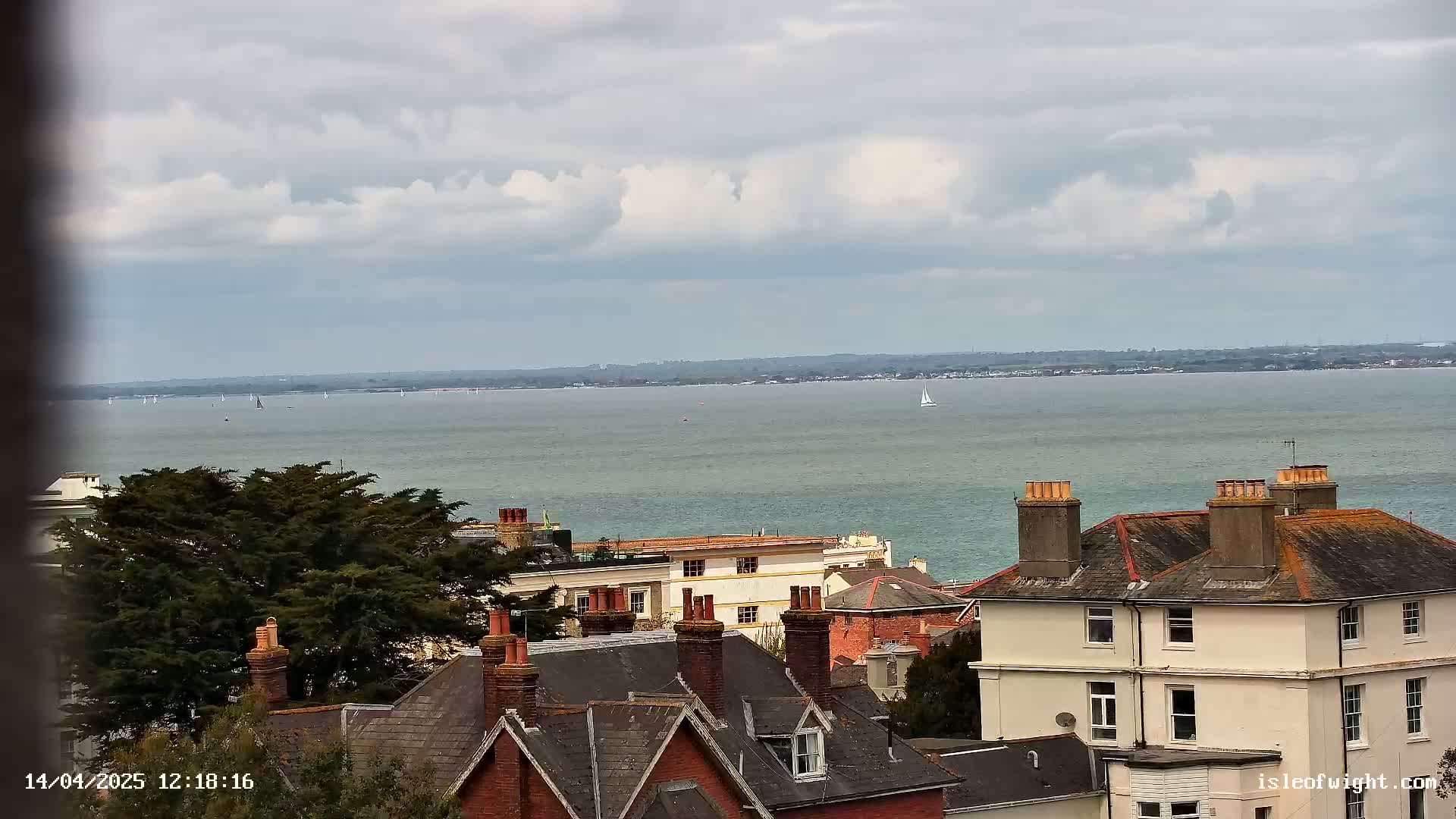 A view of a calm body of water with several sailboats, under a cloudy sky, overlooking a collection of houses with red-tiled roofs and chimneys.