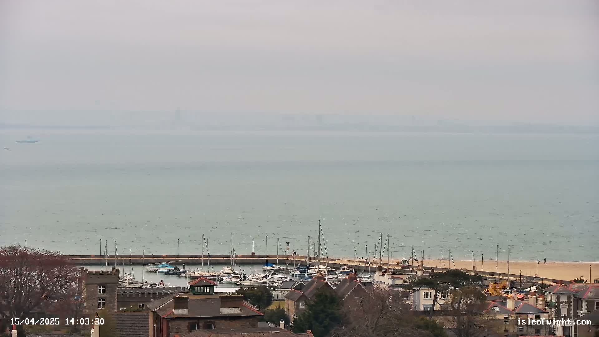 A hazy, overcast day reveals a coastal town with a marina filled with boats, a sandy beach, and houses nestled along the shoreline.
