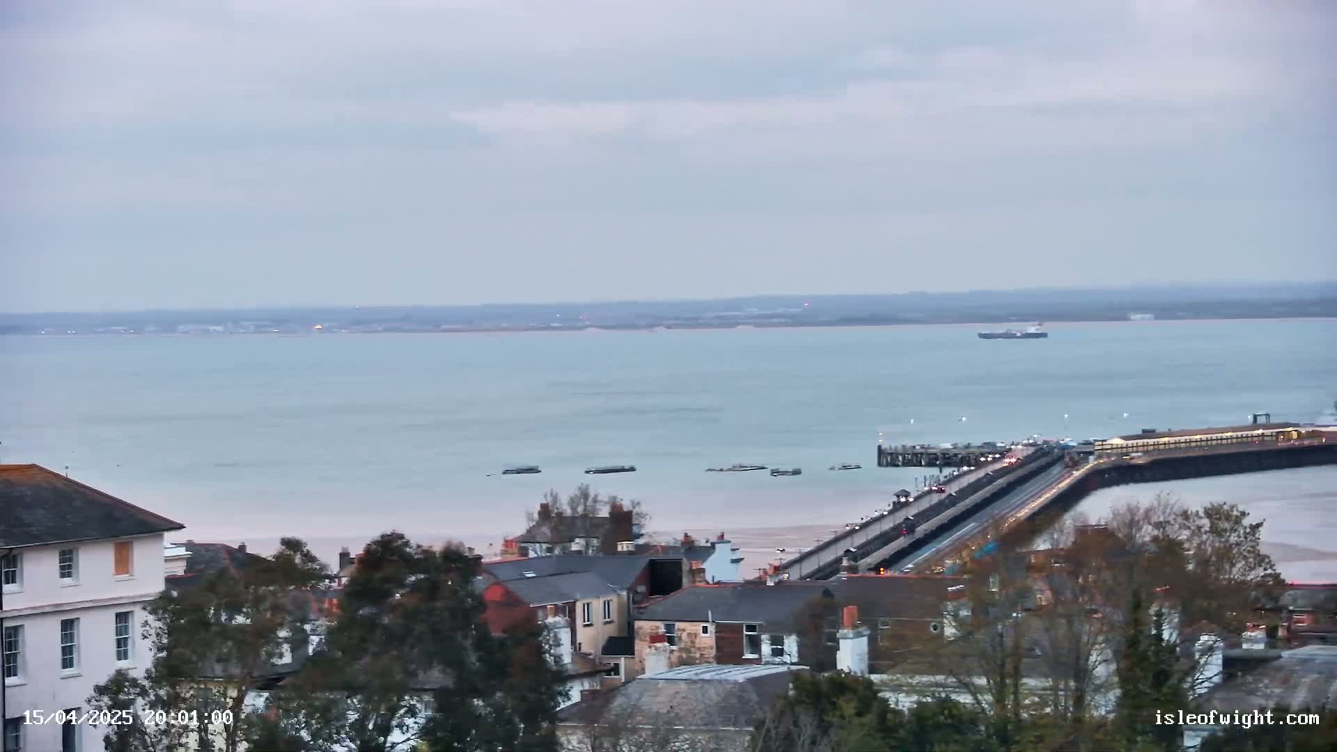 An elevated view shows a town with a long pier extending into a calm, light-blue body of water under an overcast sky.