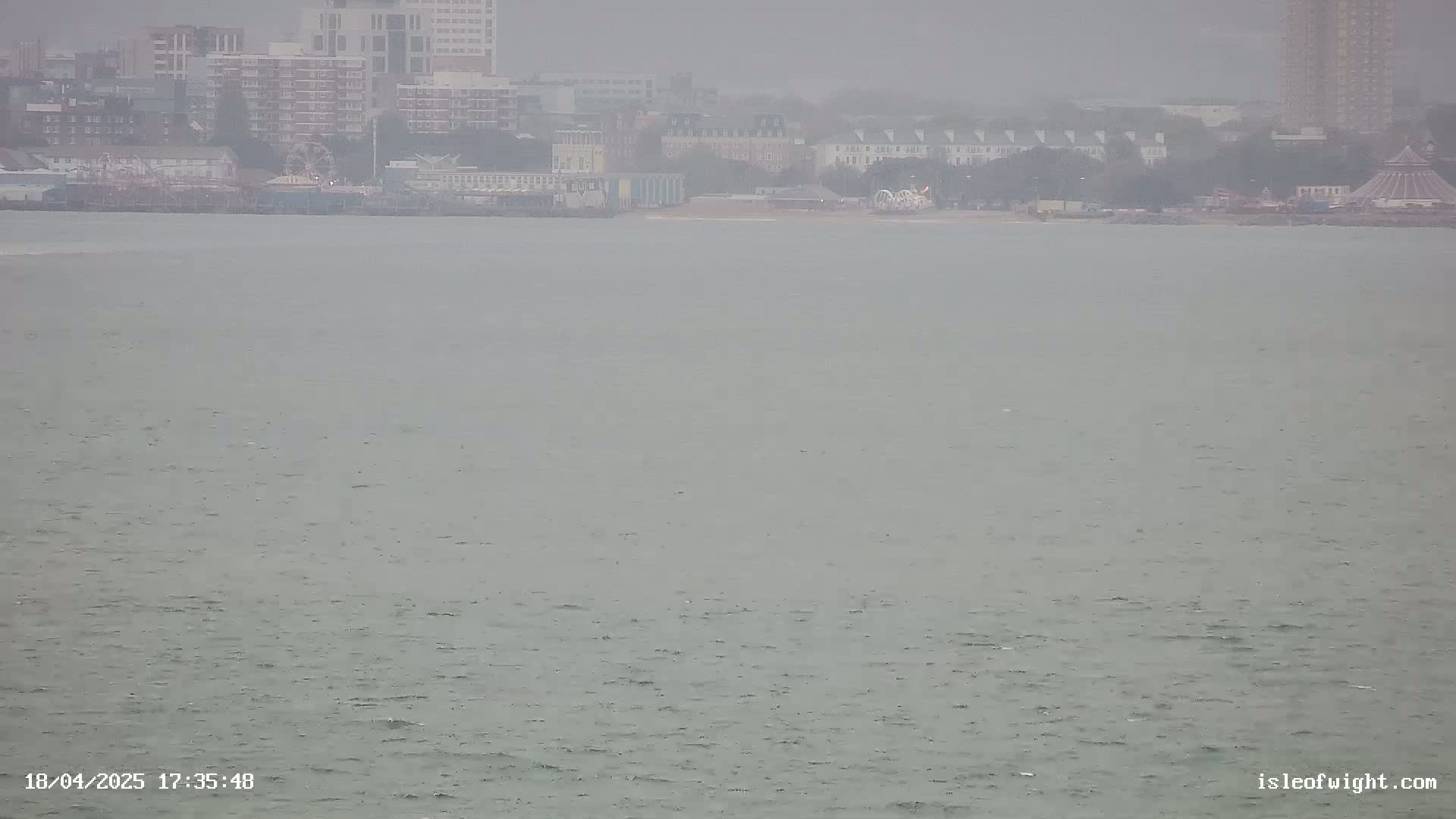 A hazy view shows a body of water with a distant shoreline featuring buildings, a Ferris wheel, and other structures under an overcast sky.