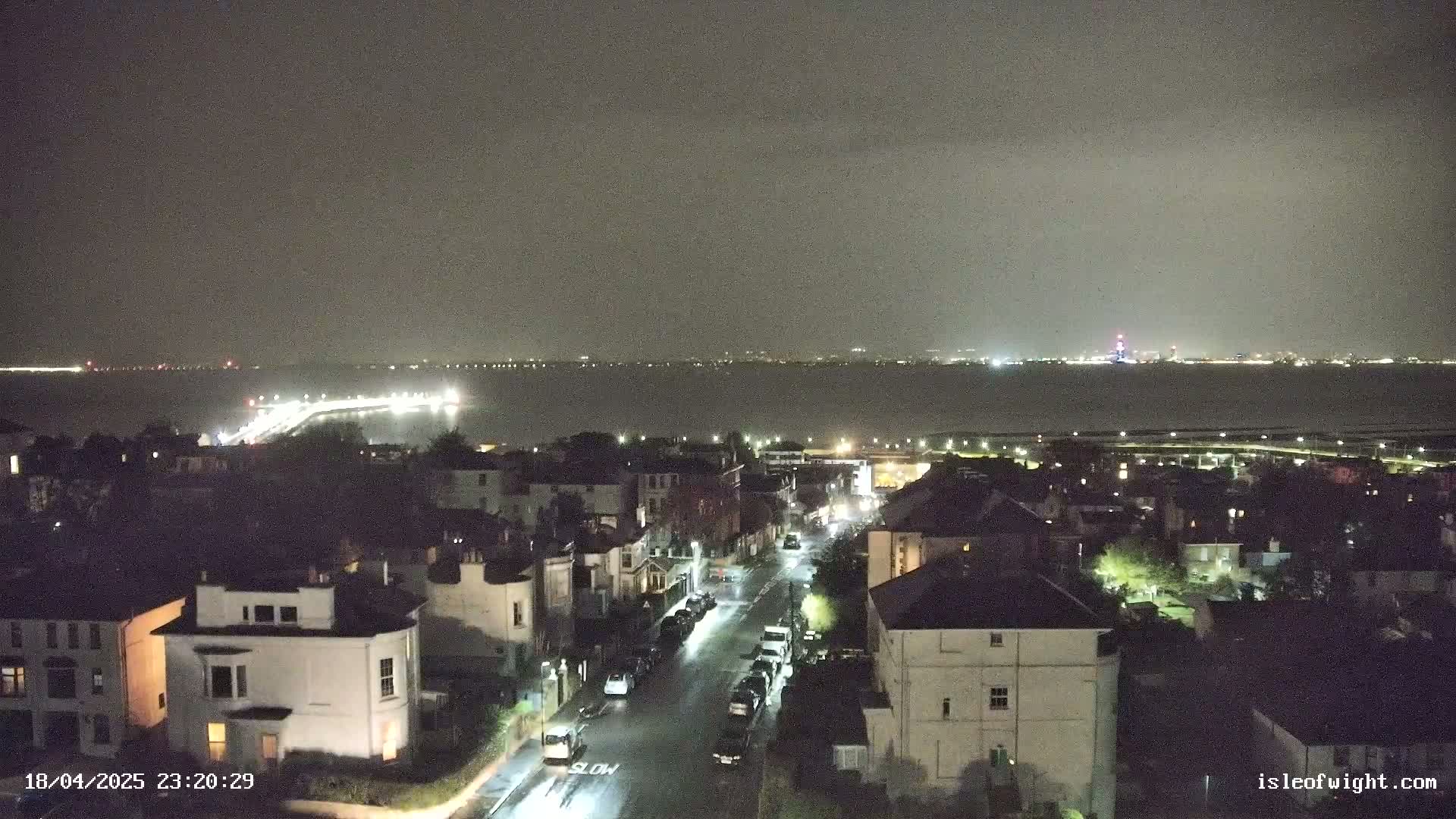 A nighttime view shows a residential street lined with houses, overlooking a body of water with a distant, illuminated city skyline under a dark, cloudy sky.