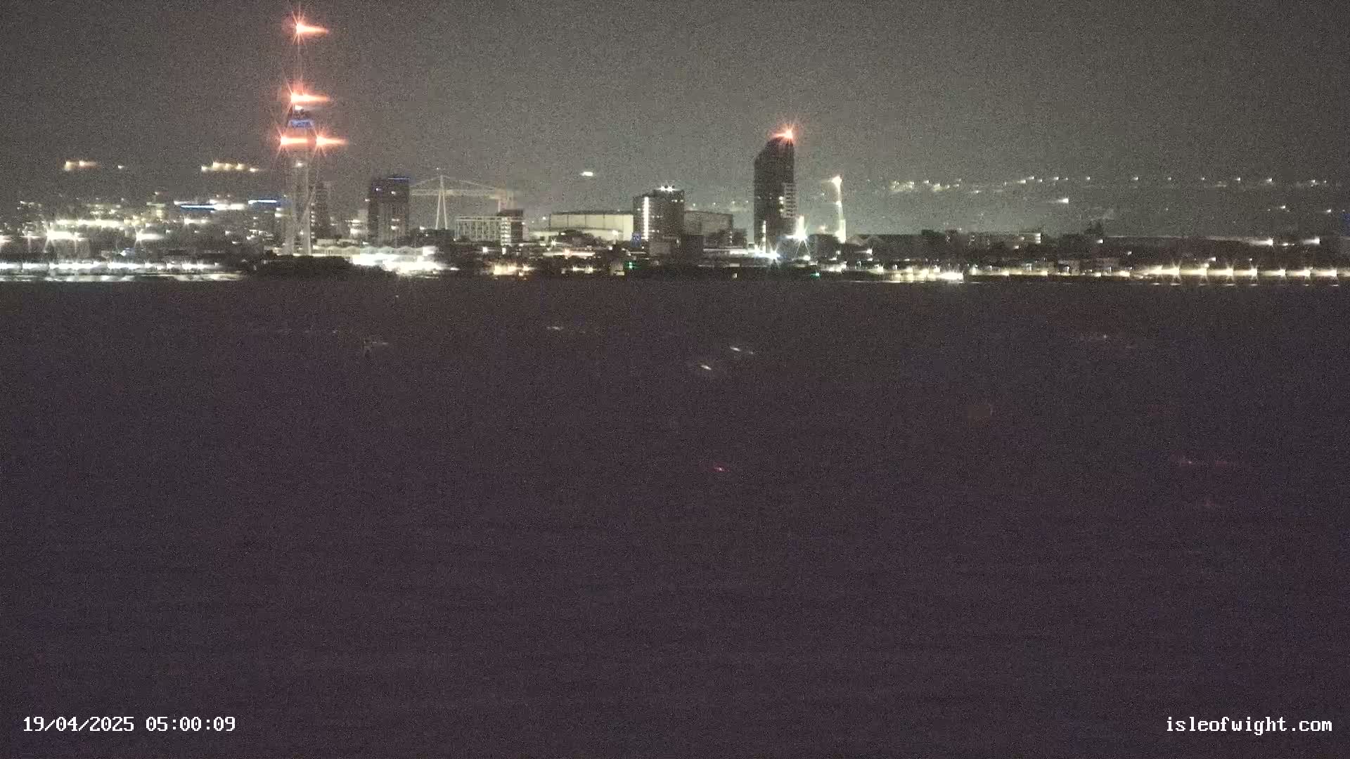 A nighttime view of a city skyline across a dark body of water, with several tall buildings and bright lights visible.