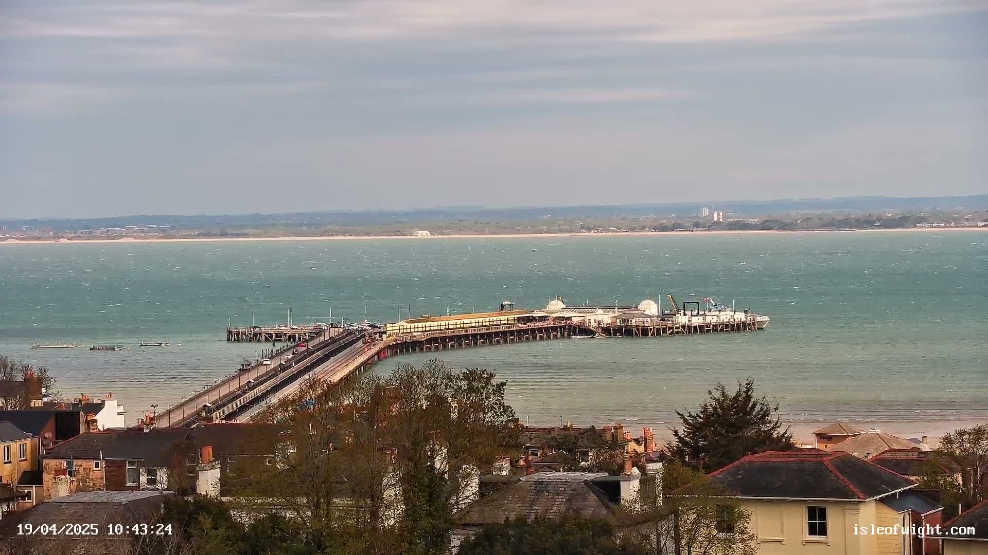 A long pier extends from a town into a choppy, greenish-blue sea under a cloudy sky.
