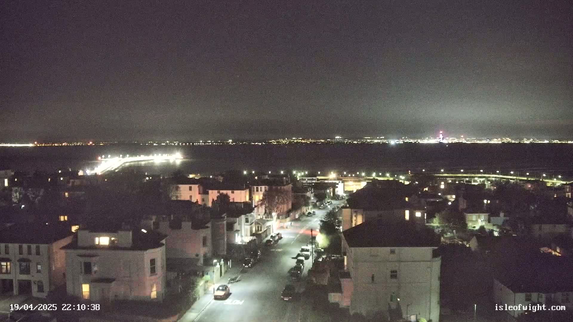 A nighttime, high-angle view shows a residential street with parked cars and a body of water with city lights in the distance under a clear, dark sky.