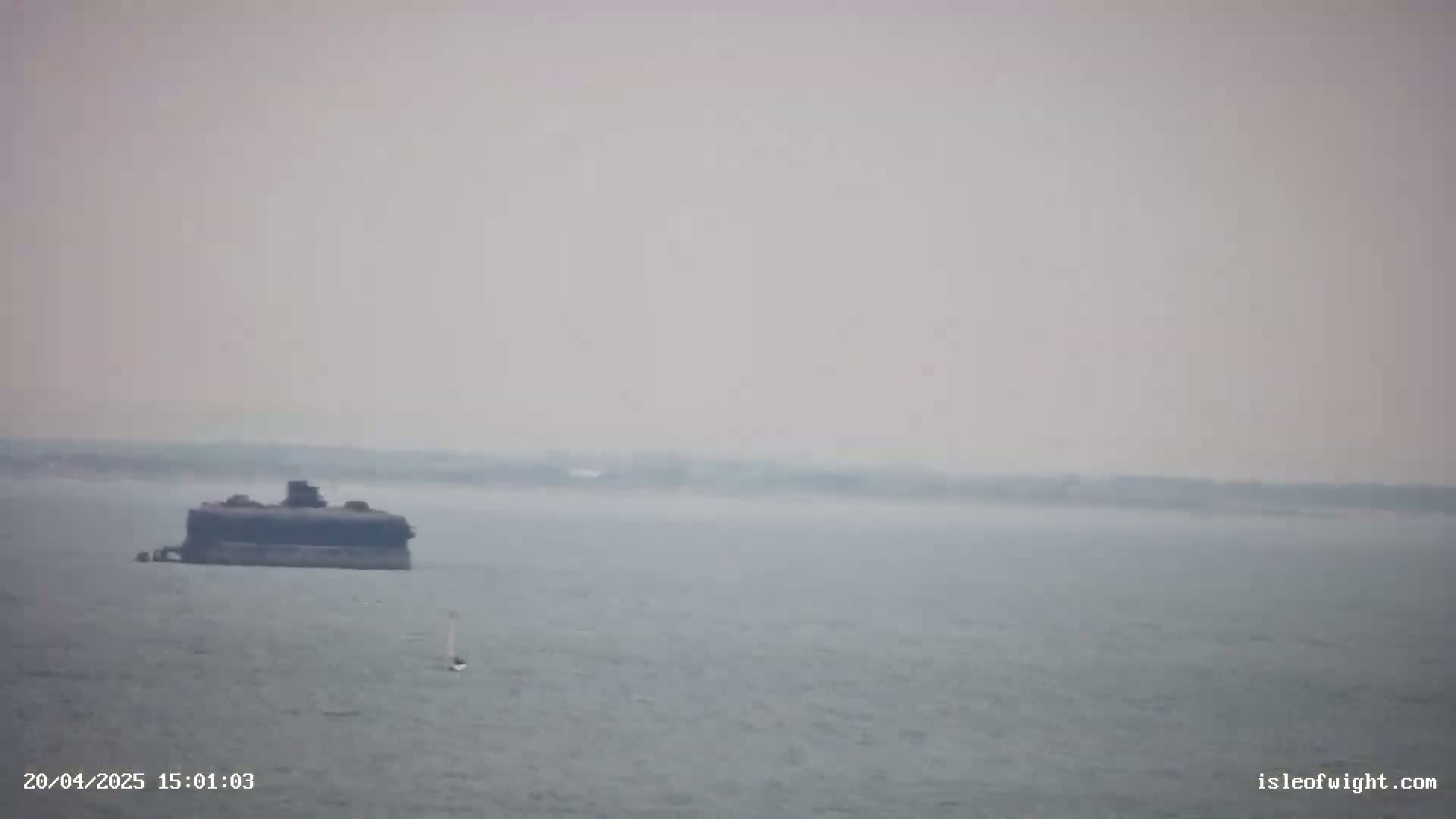 A large, dark structure sits in calm, hazy water, with a small sailboat visible in the distance.