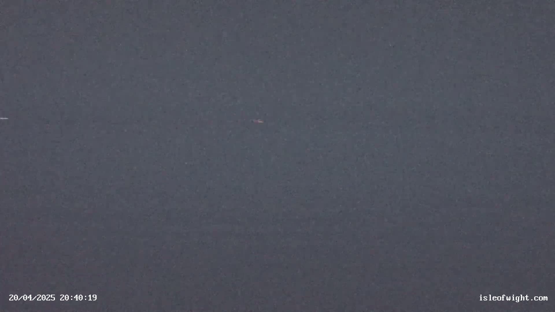 A dark gray, possibly nighttime, view of a calm body of water with a few tiny, indistinct objects visible on the surface.