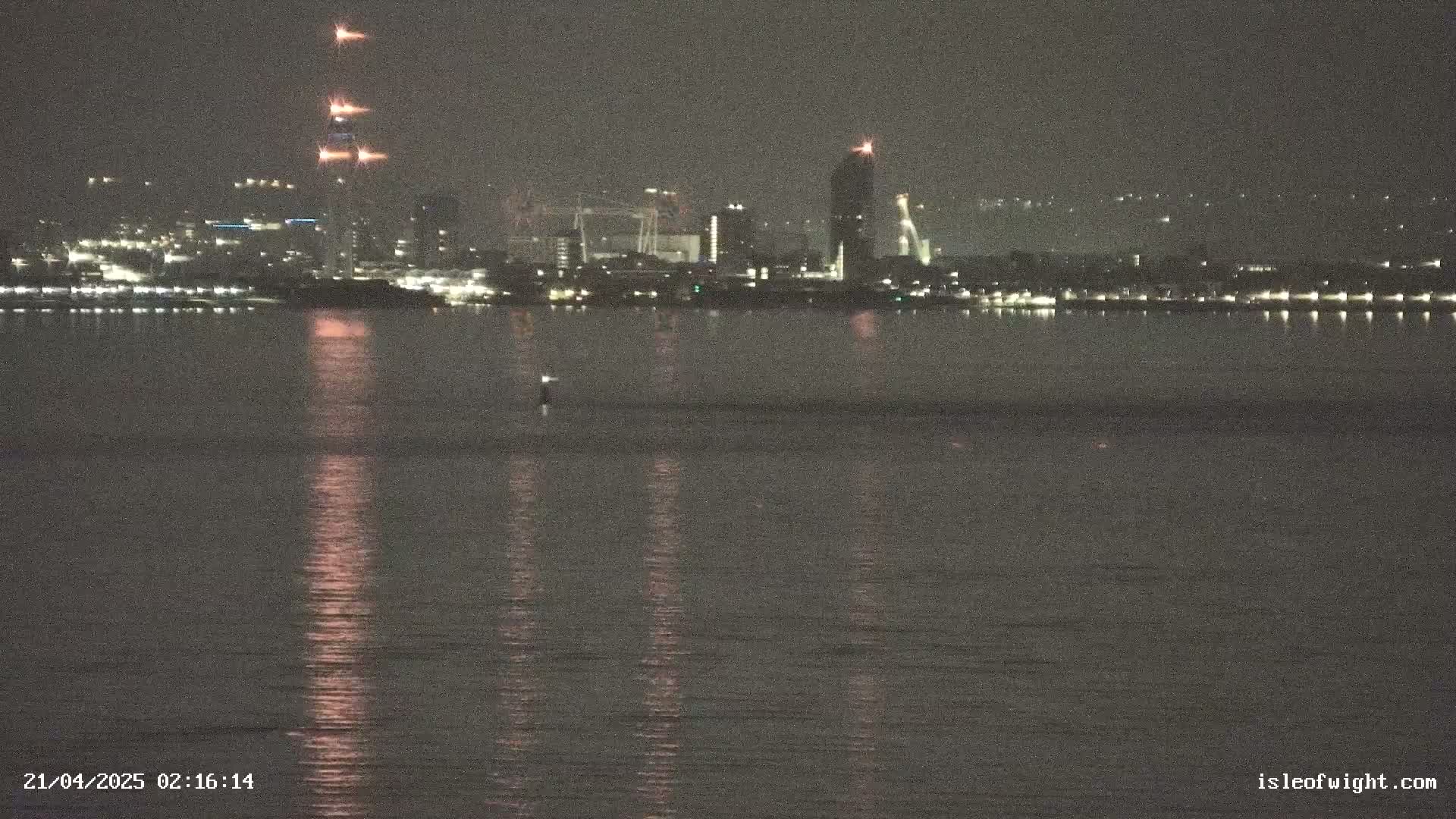A nighttime view across a calm body of water shows a city skyline with several tall buildings and construction lights.