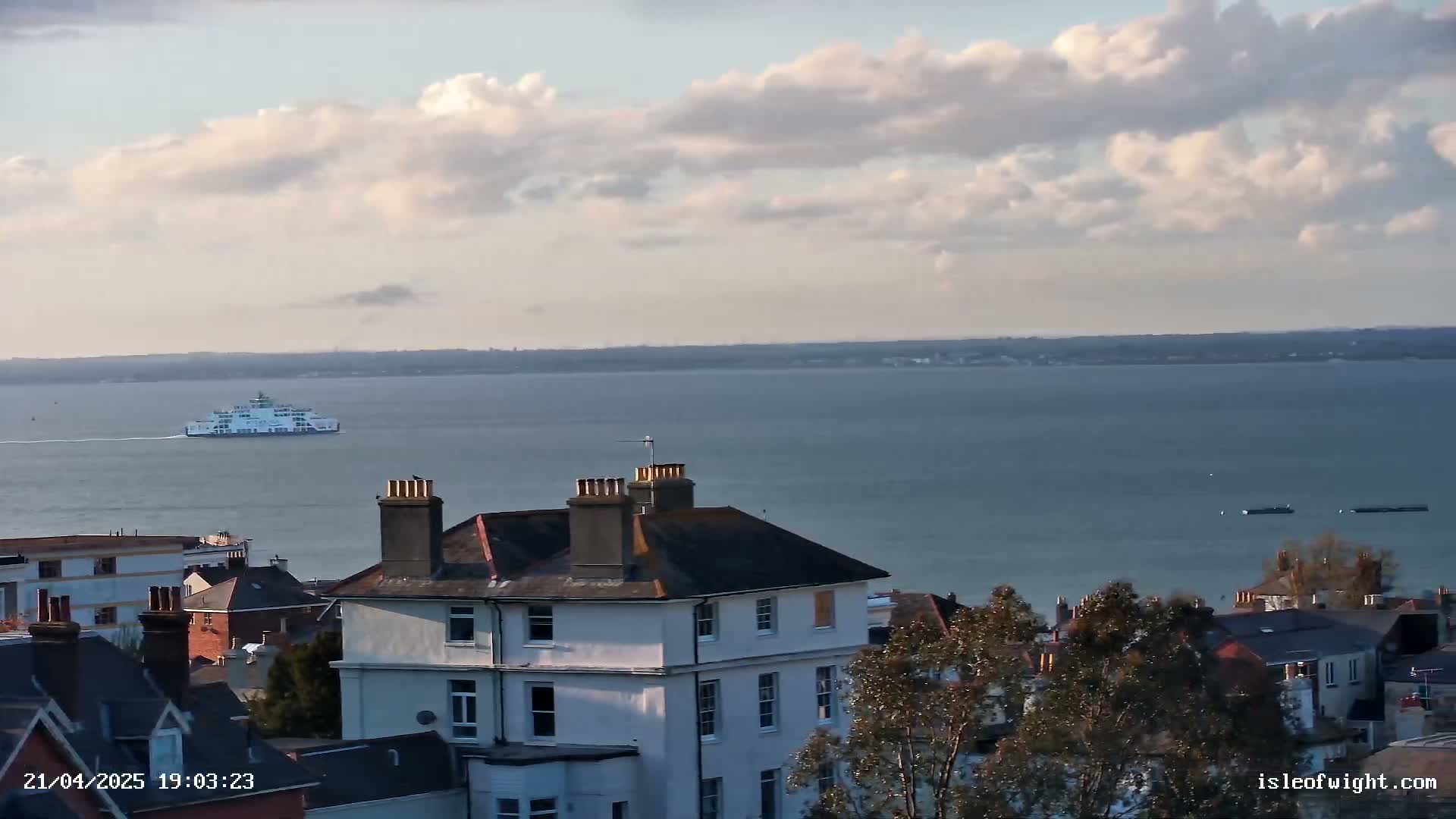A ferry sails on a calm body of water under a partly cloudy sky, viewed from an elevated position overlooking a collection of houses and trees.