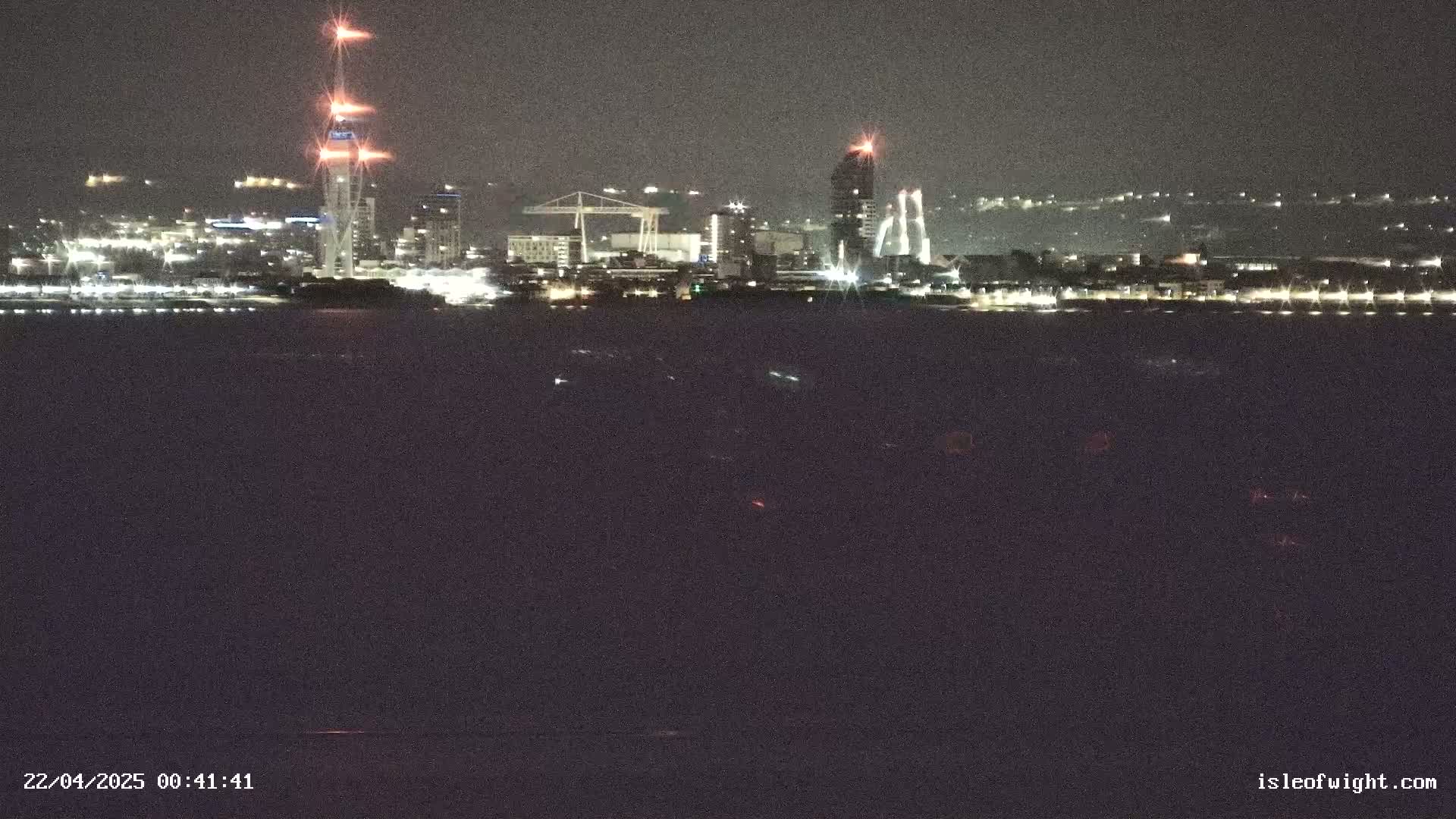 A nighttime view of a city skyline across a dark body of water, featuring several tall buildings and industrial structures illuminated.