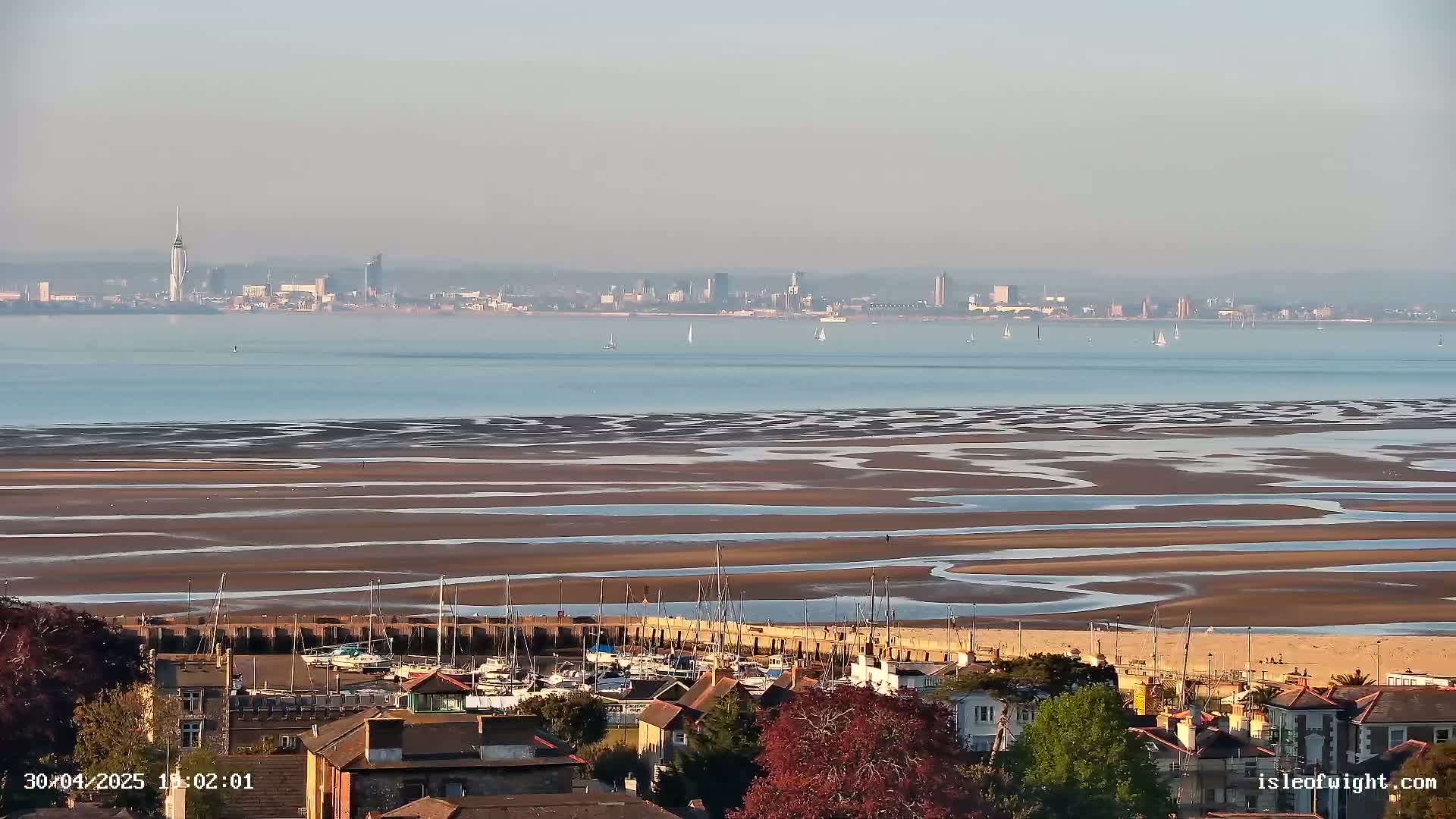 A hazy, sunny day reveals a city skyline across a wide expanse of calm water and exposed, patterned mudflats at low tide, with a small harbor and residential area in the foreground.