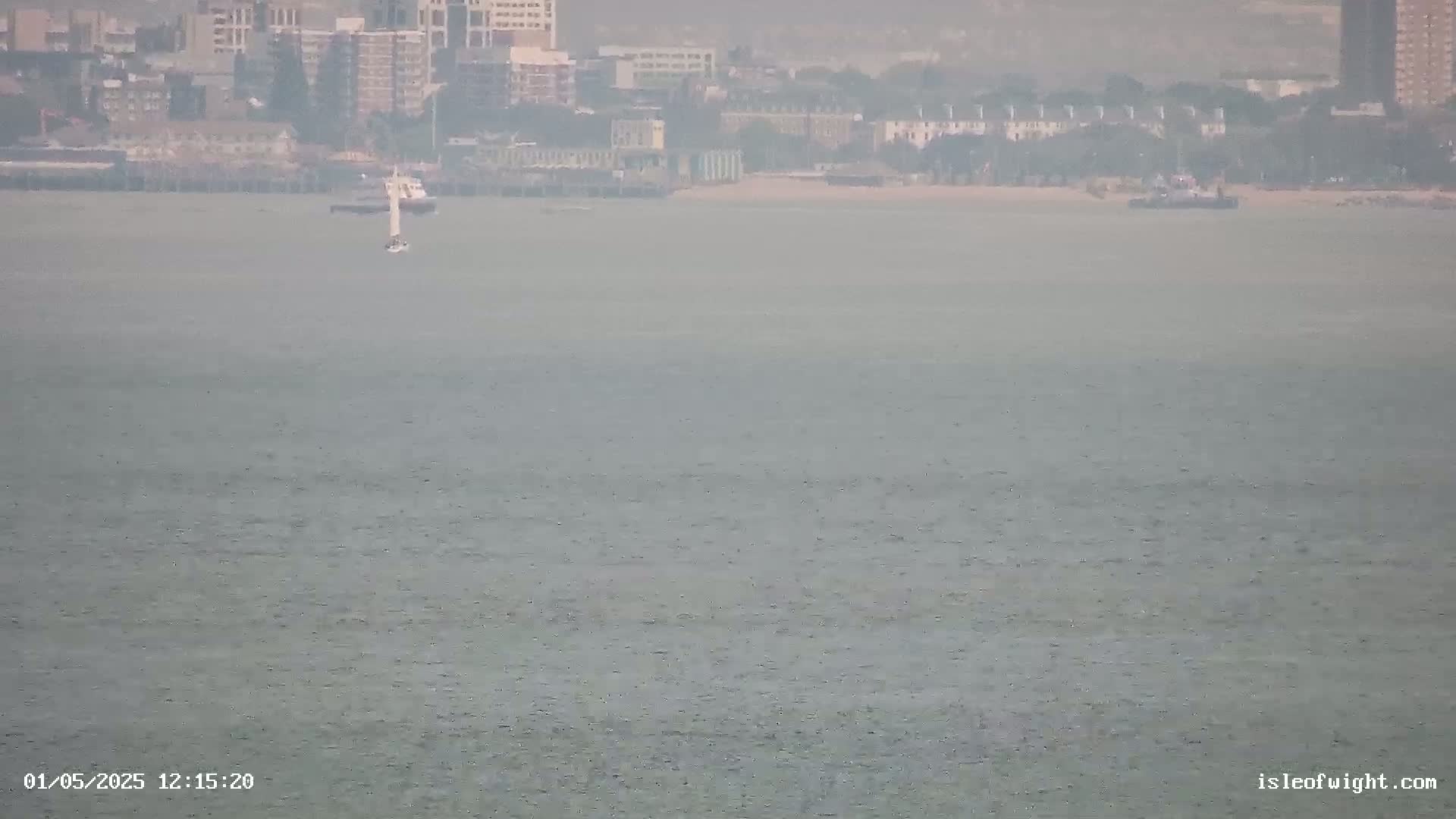 A hazy view of a body of water shows a sailboat and a distant shoreline with buildings under an overcast sky.