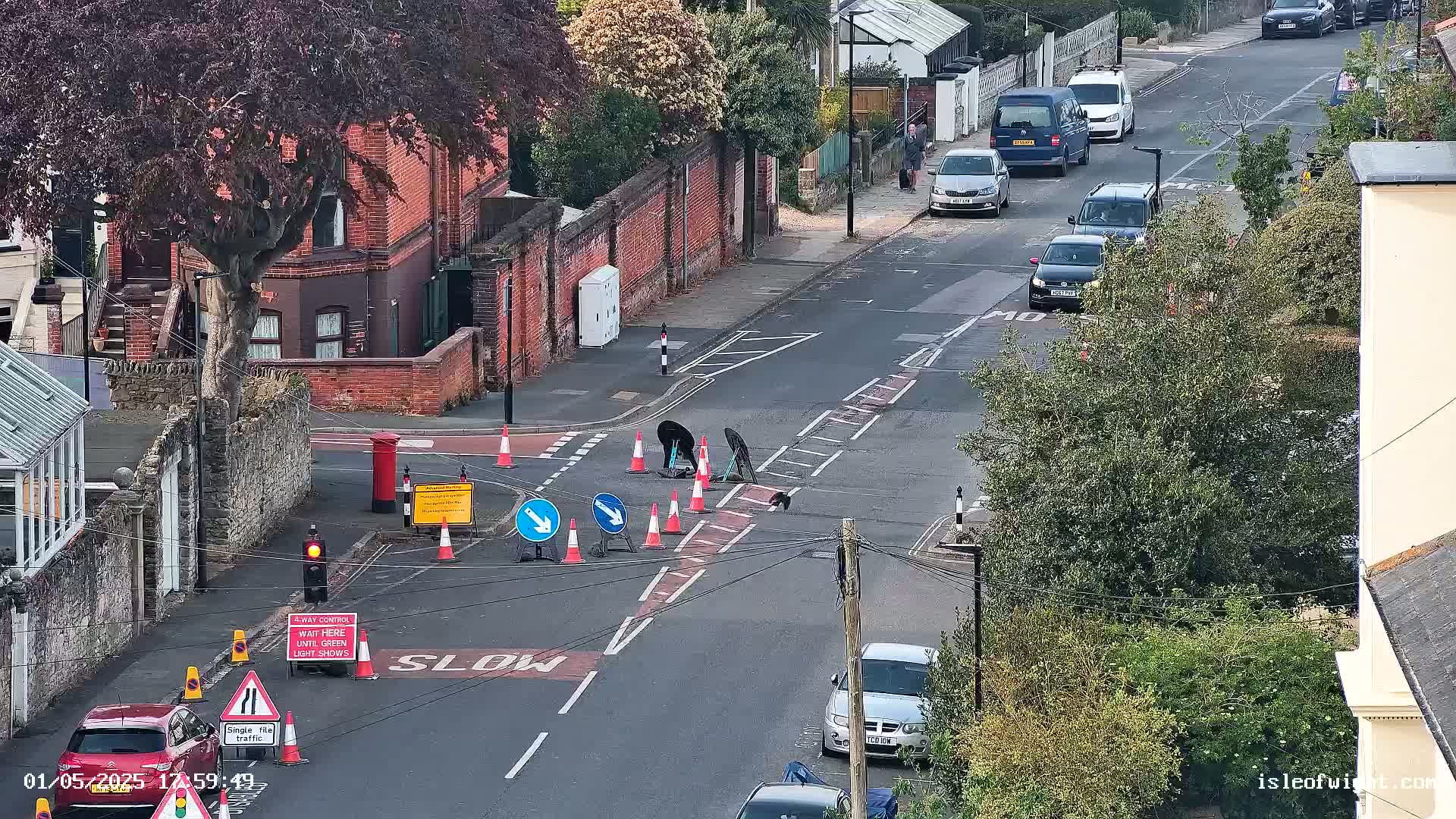 Road workers are repairing a section of road at a street intersection surrounded by parked cars and houses, under a partly sunny sky.