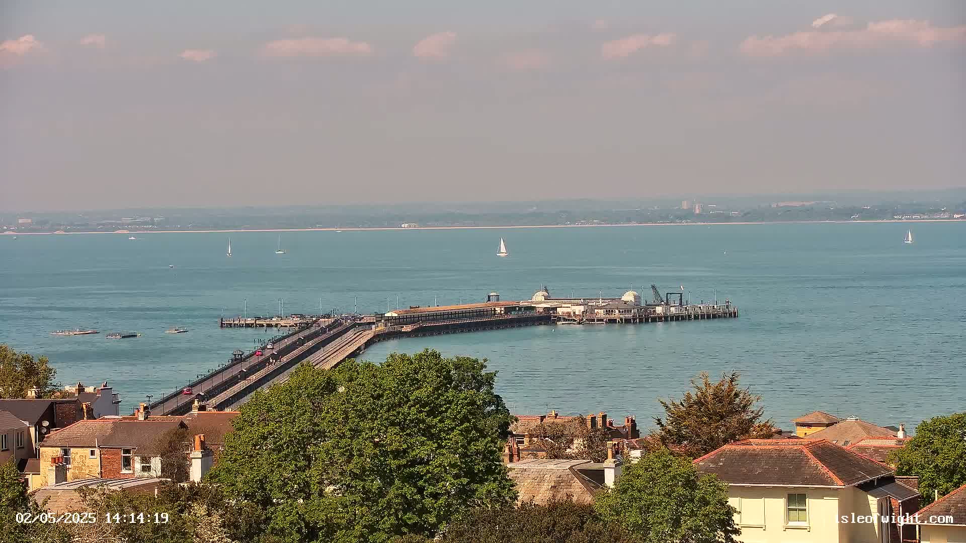 A long pier extends into a calm, blue body of water under a partly cloudy sky, with a collection of buildings and green trees visible in the foreground.