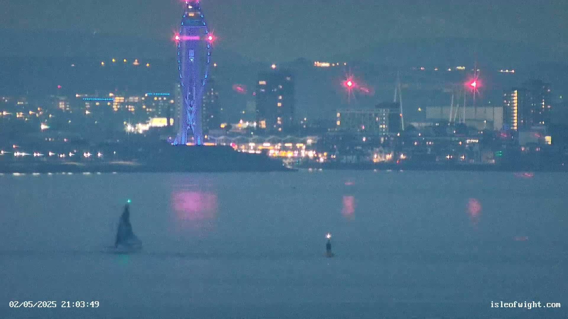 A nighttime view of a city skyline with a tall, illuminated tower across a calm body of water, with two small boats visible.