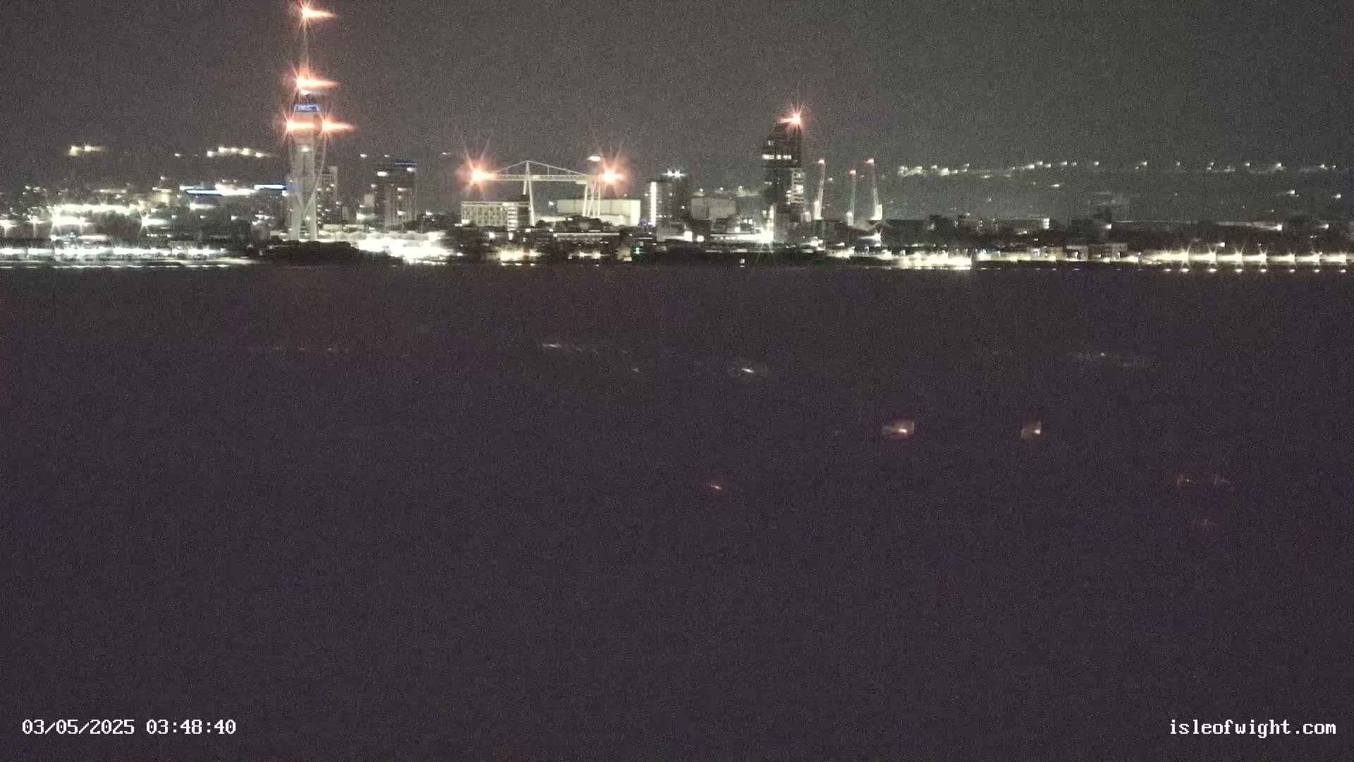 A nighttime view of a city skyline across a dark body of water, illuminated by the lights of buildings and what appears to be construction activity.
