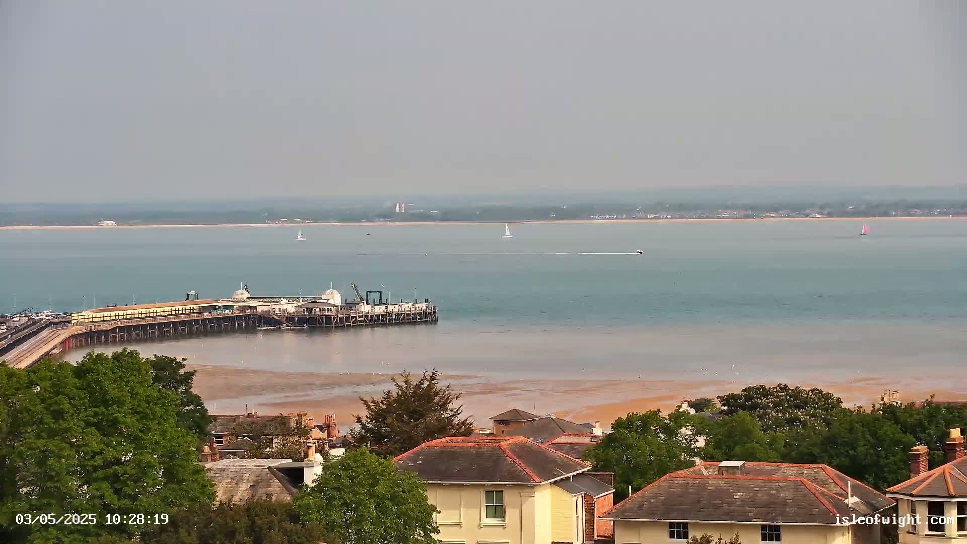 A long pier extends into a calm, hazy body of water, with a town and several sailboats visible in the distance.