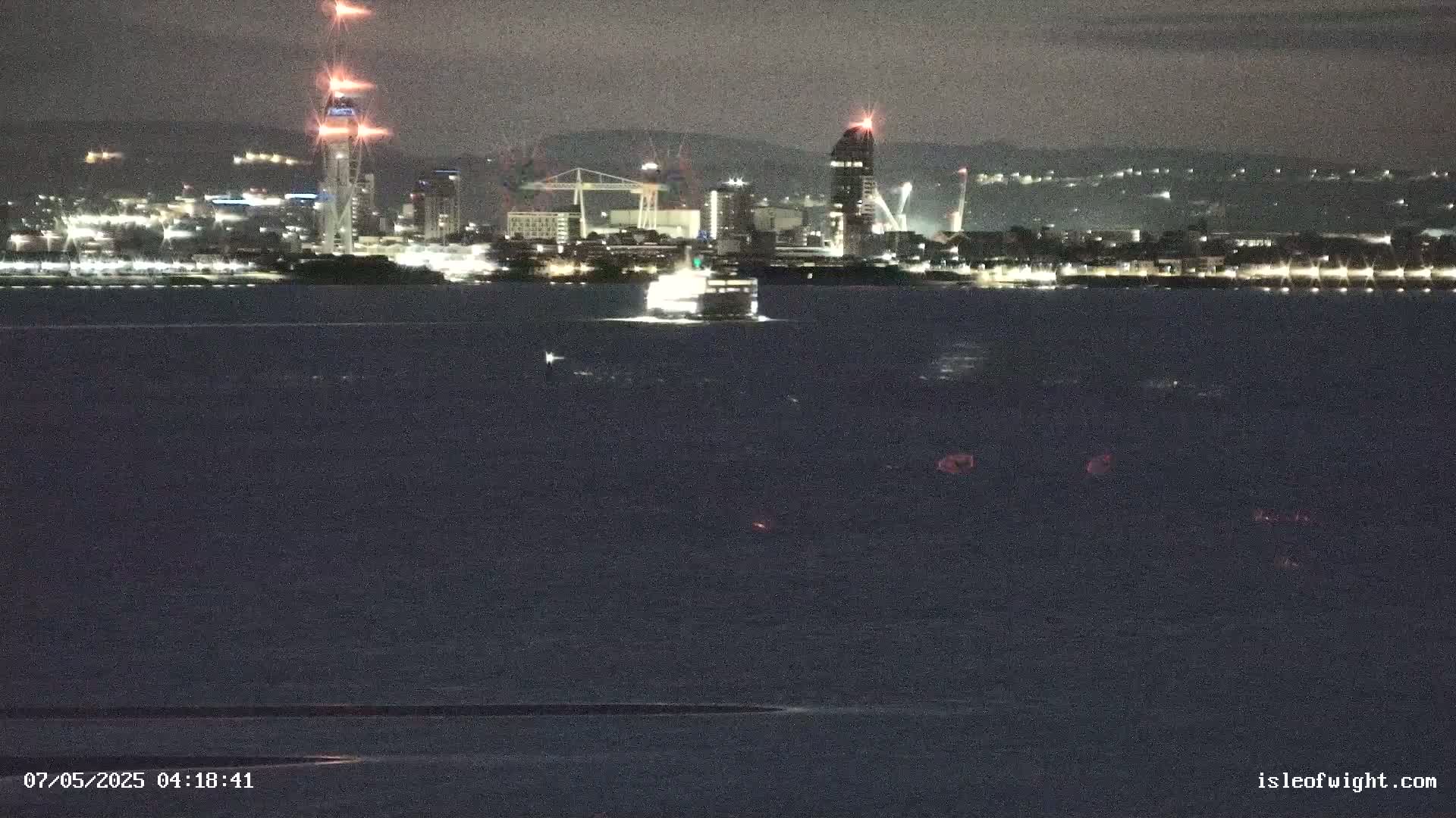 A nighttime view across dark water shows a ferry approaching a brightly lit city skyline under a clear, dark sky.