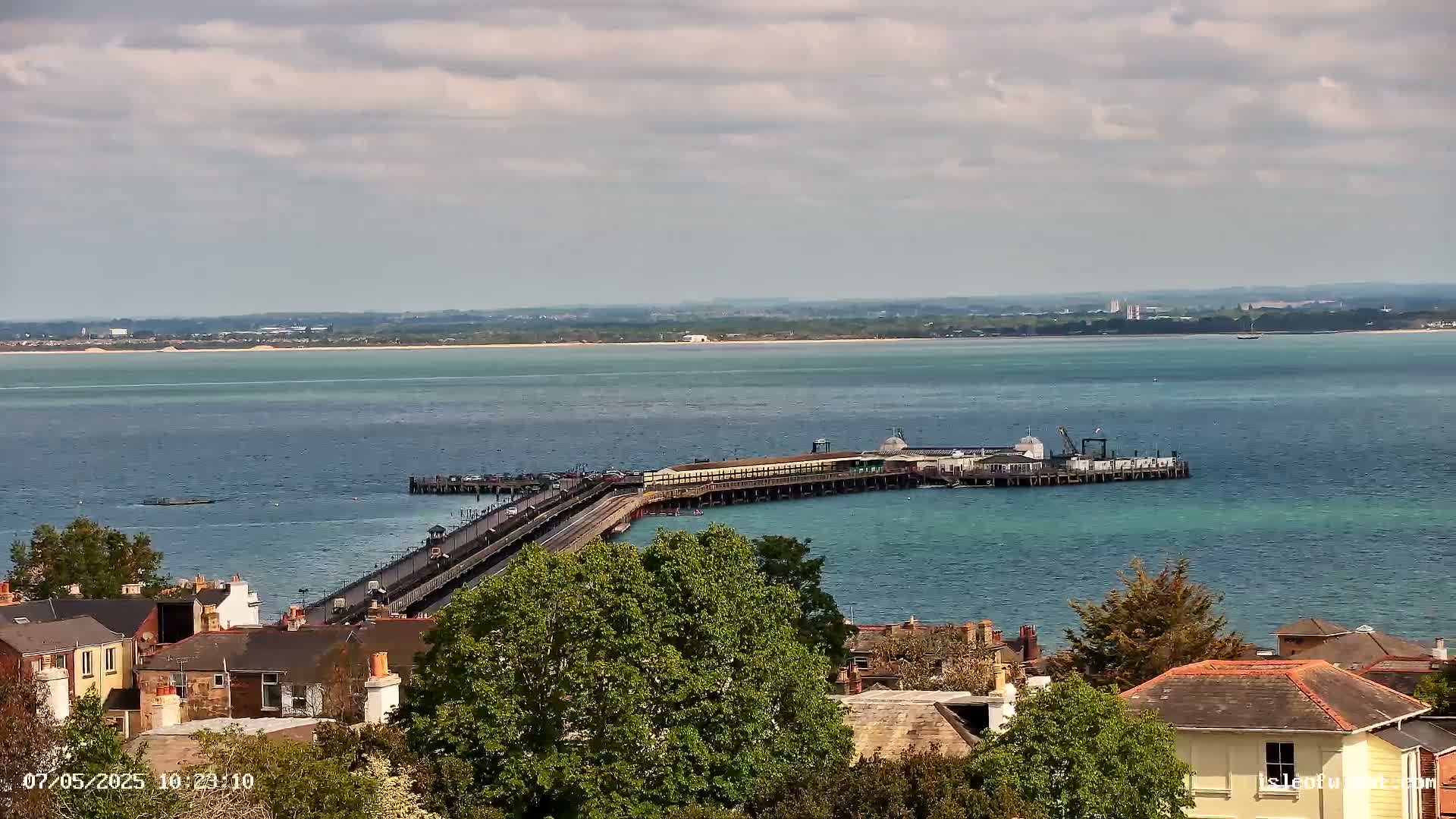 A long pier extends into a calm, blue-green body of water under a partly cloudy sky, with residential buildings and lush green trees visible in the foreground.