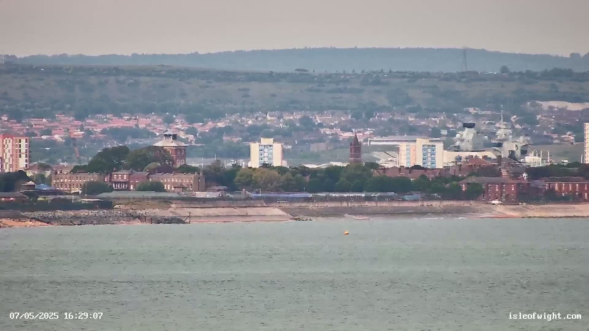 A distant view of a coastal town with several buildings, including a brick structure with a tower, under a hazy sky over calm water.