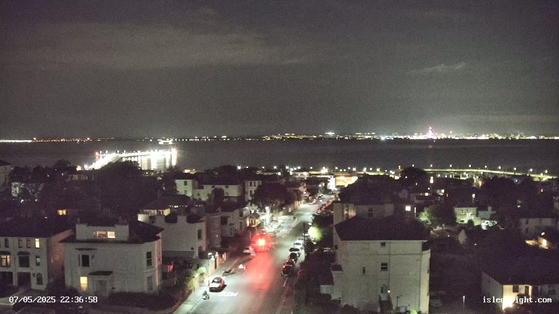 A nighttime, high-angle view shows a residential street with parked cars and a pier extending into a dark body of water with a distant city skyline visible under a cloudy sky.