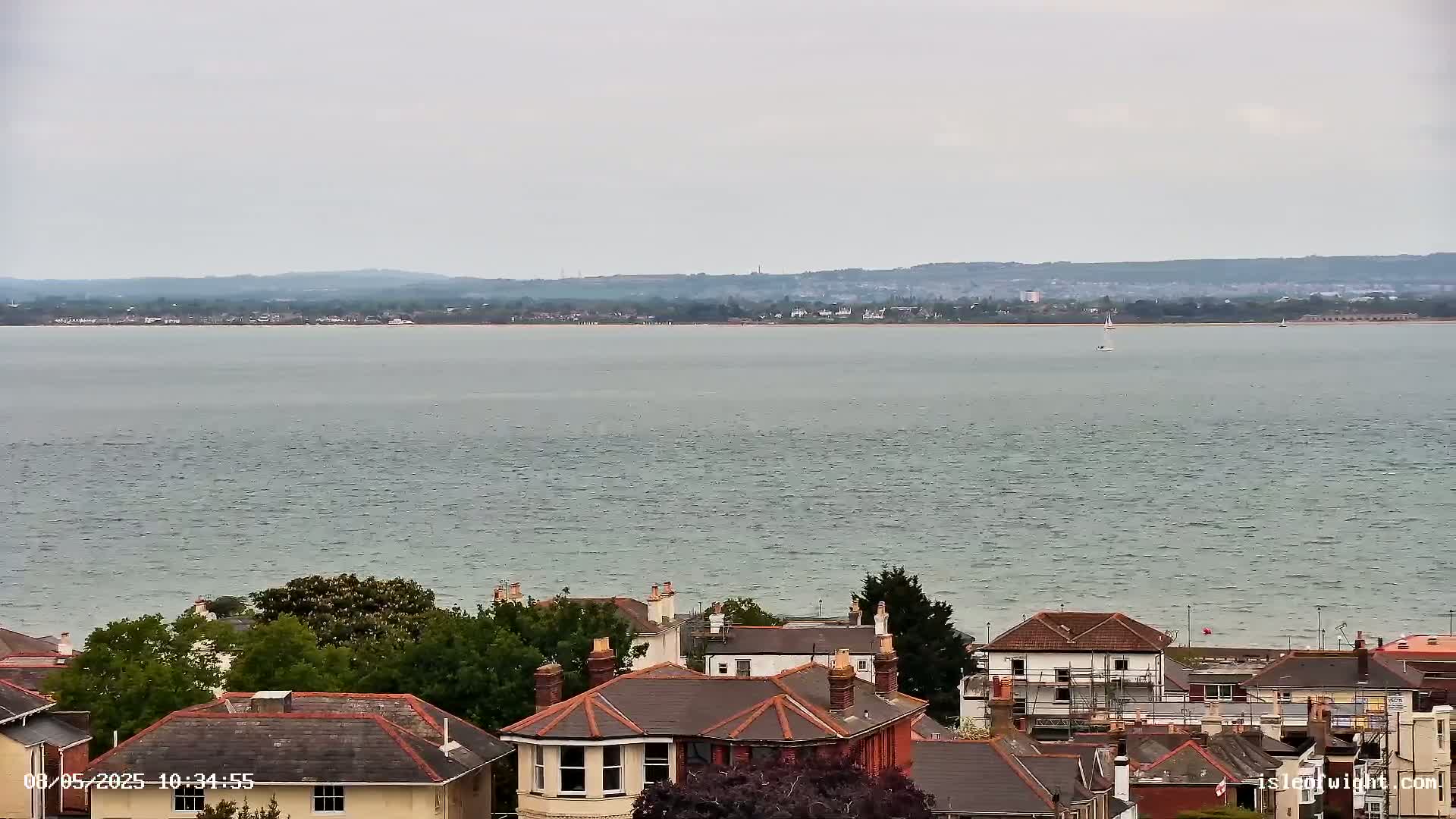 A view of a calm body of water with a distant shoreline and a collection of houses with brown roofs nestled amongst green trees in the foreground under an overcast sky.