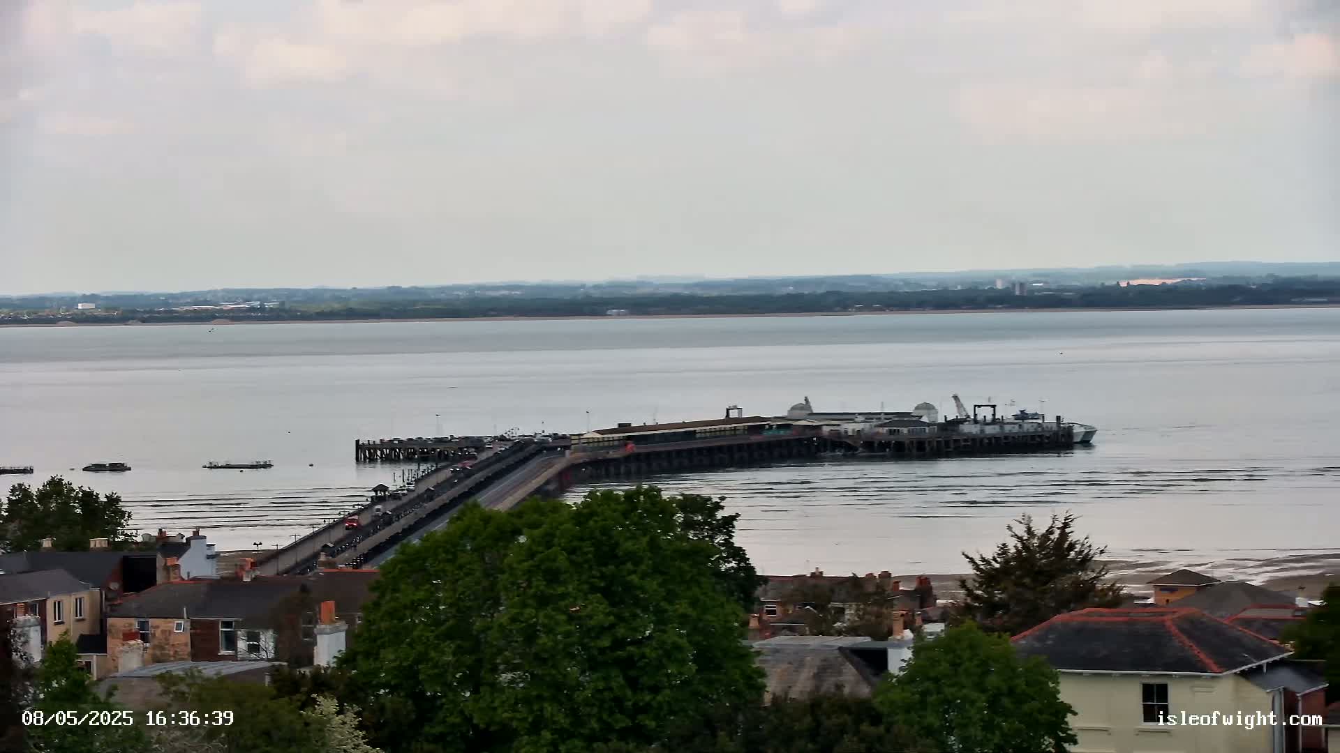 A long pier extends into a calm, wide body of water under an overcast sky, with a town visible in the foreground and a distant shoreline on the horizon.