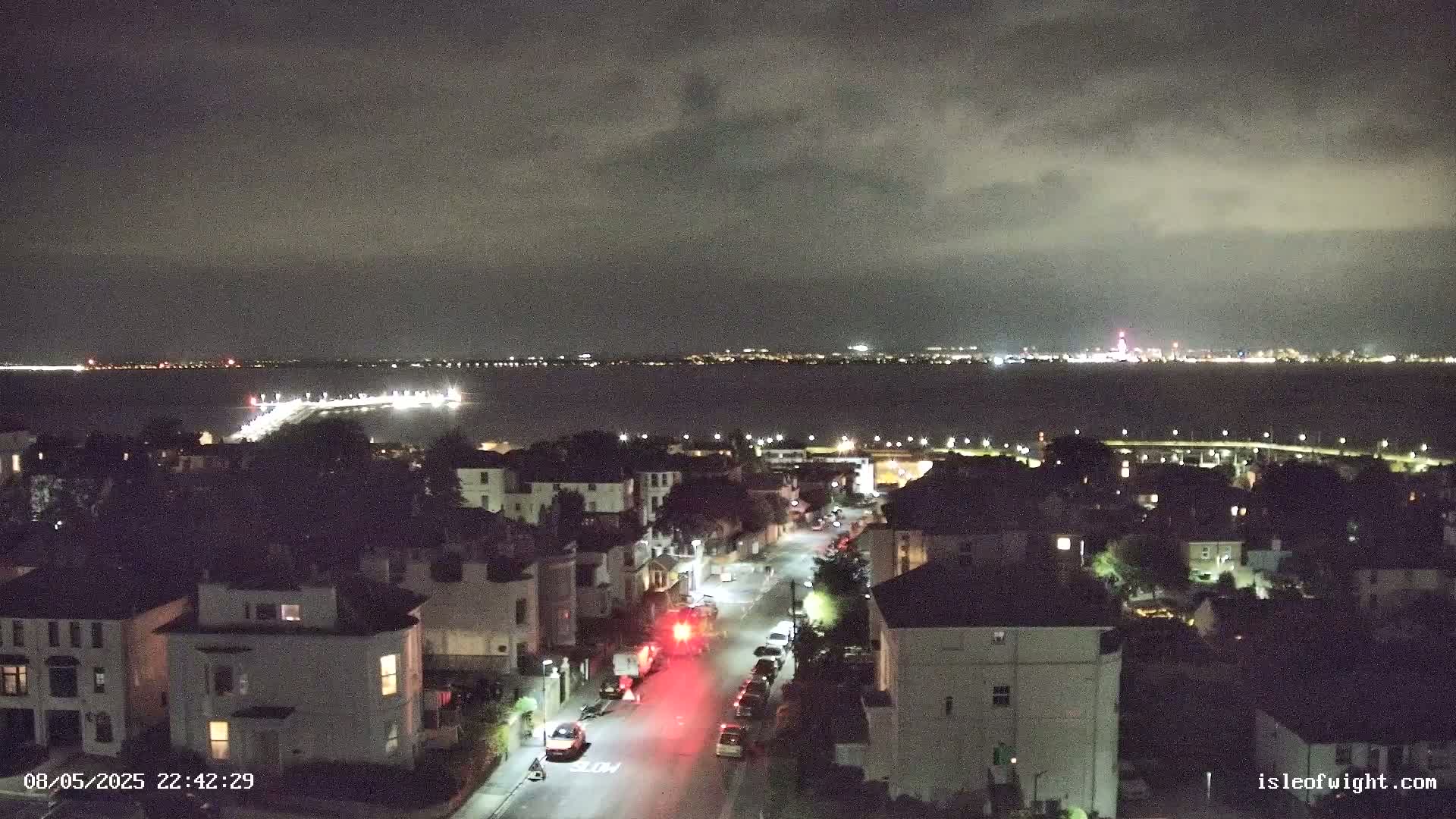 A nighttime view of a residential street overlooking a body of water with a distant city skyline under a cloudy sky.