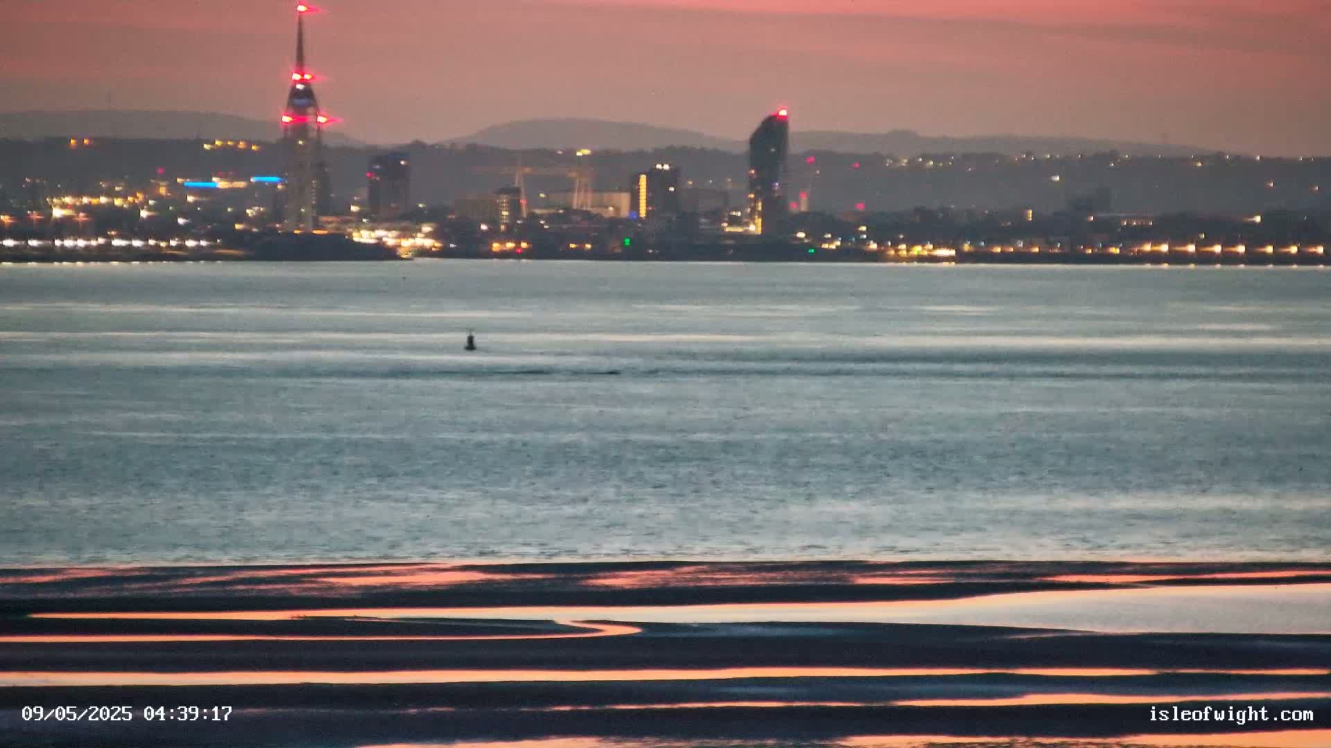 A city skyline with illuminated buildings and a tall tower is visible across a calm body of water reflecting the pink and orange hues of the twilight sky, while the foreground shows a flat, sandy expanse with channels of water reflecting the same colors.