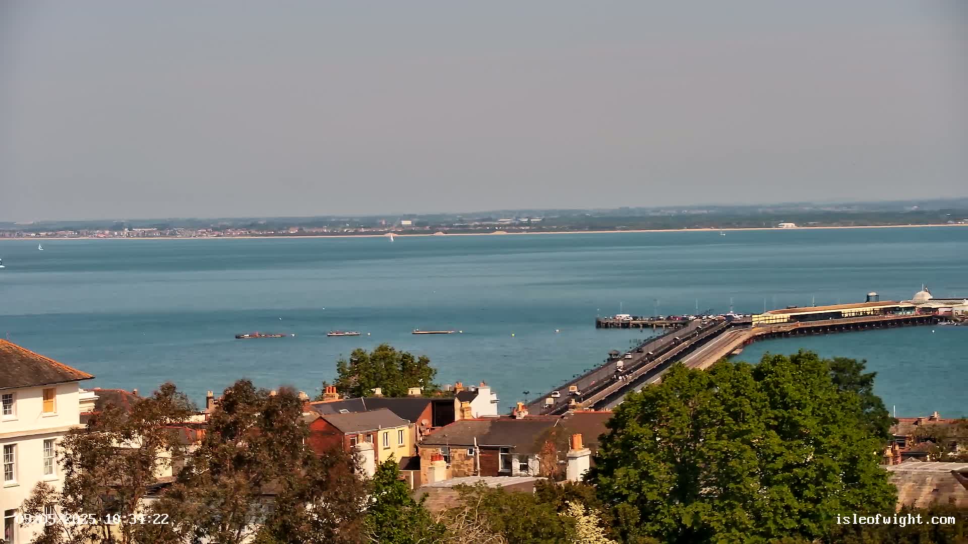 A long pier extends from a town into a calm, blue body of water on a bright, sunny day, with a distant shoreline visible across the water.