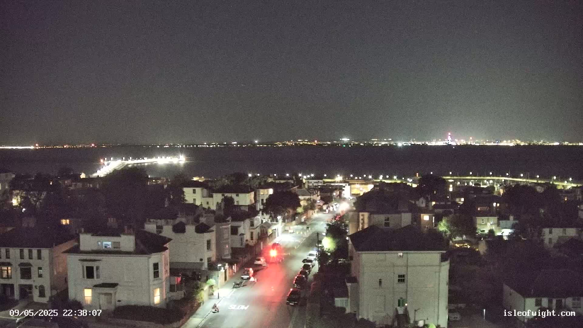 A nighttime aerial view shows a residential street with parked cars and a few moving vehicles, overlooking a body of water with a distant city skyline illuminated under a clear, dark sky.