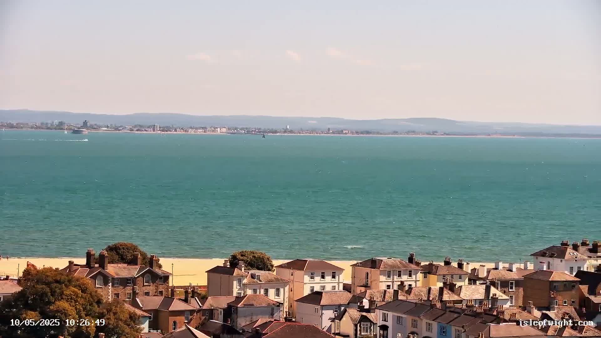 A coastal town with houses and a sandy beach overlooks a calm, teal-colored sea on a sunny day.