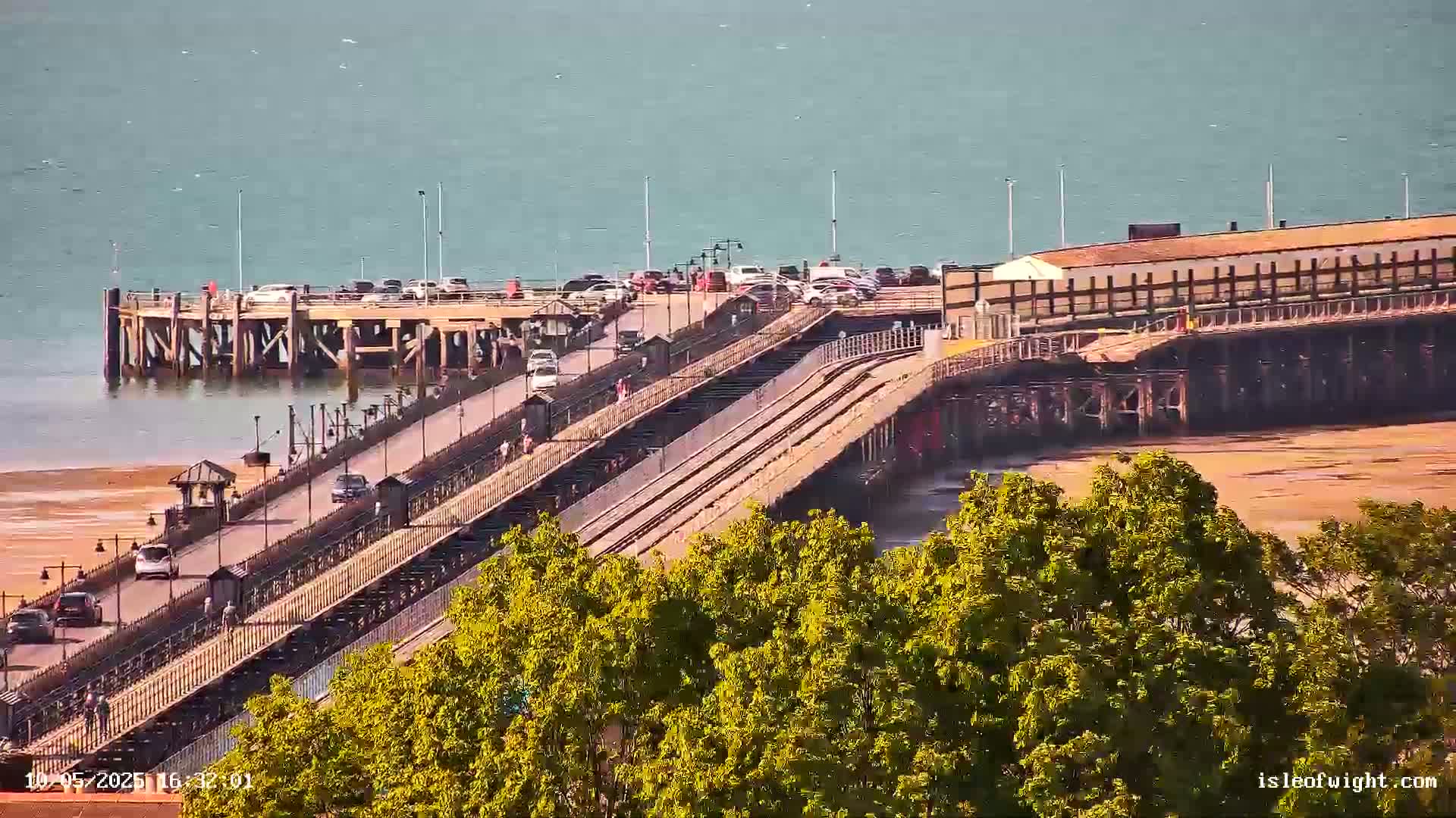 A long pier extends over calm, light-blue water, with cars driving along it and a sandy beach visible at low tide, partially obscured by green trees in the foreground.