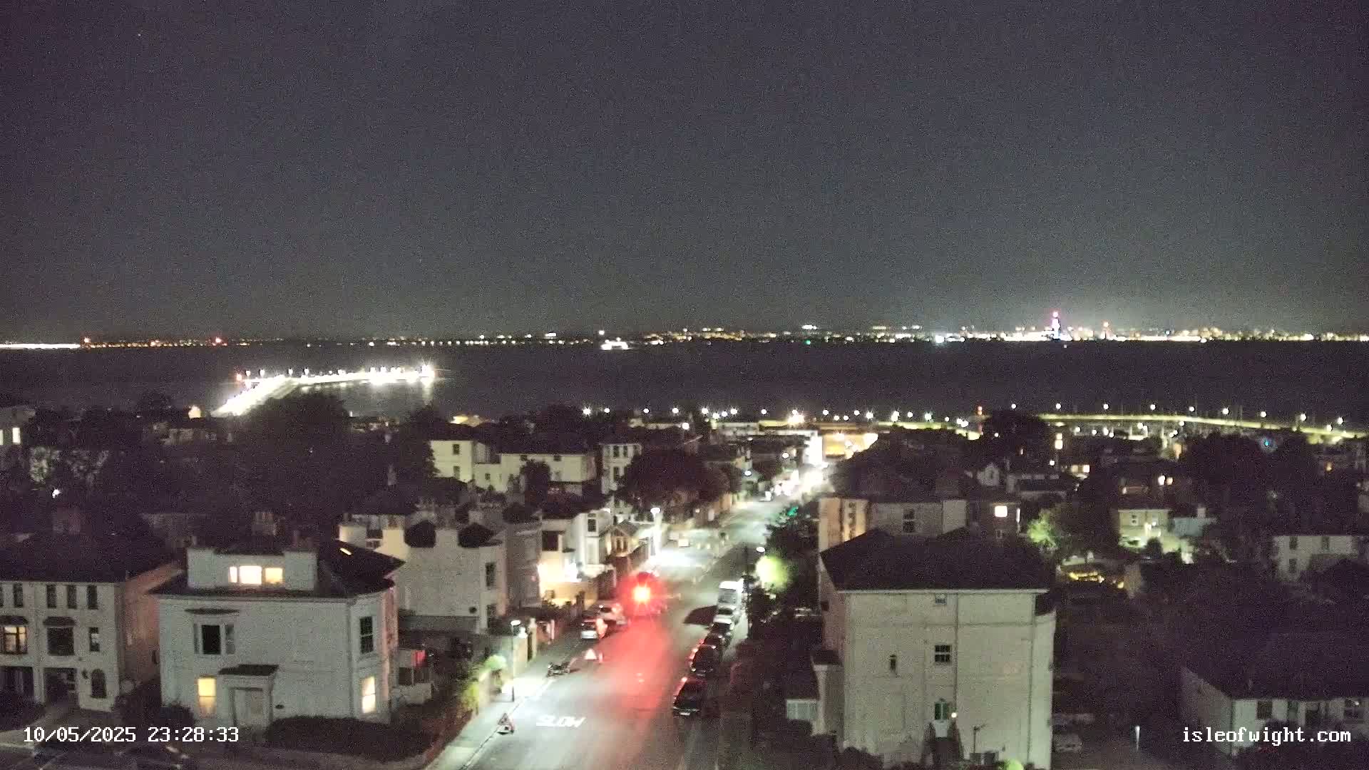 A nighttime, high-angle view shows a residential street with cars and houses illuminated, overlooking a calm body of water with a distant city skyline under a clear, dark sky.