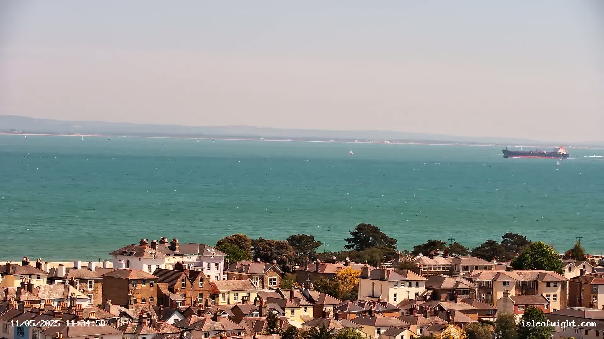 A coastal town with many houses is situated overlooking a teal-colored sea on a sunny day, with a large cargo ship and several sailboats visible in the distance.