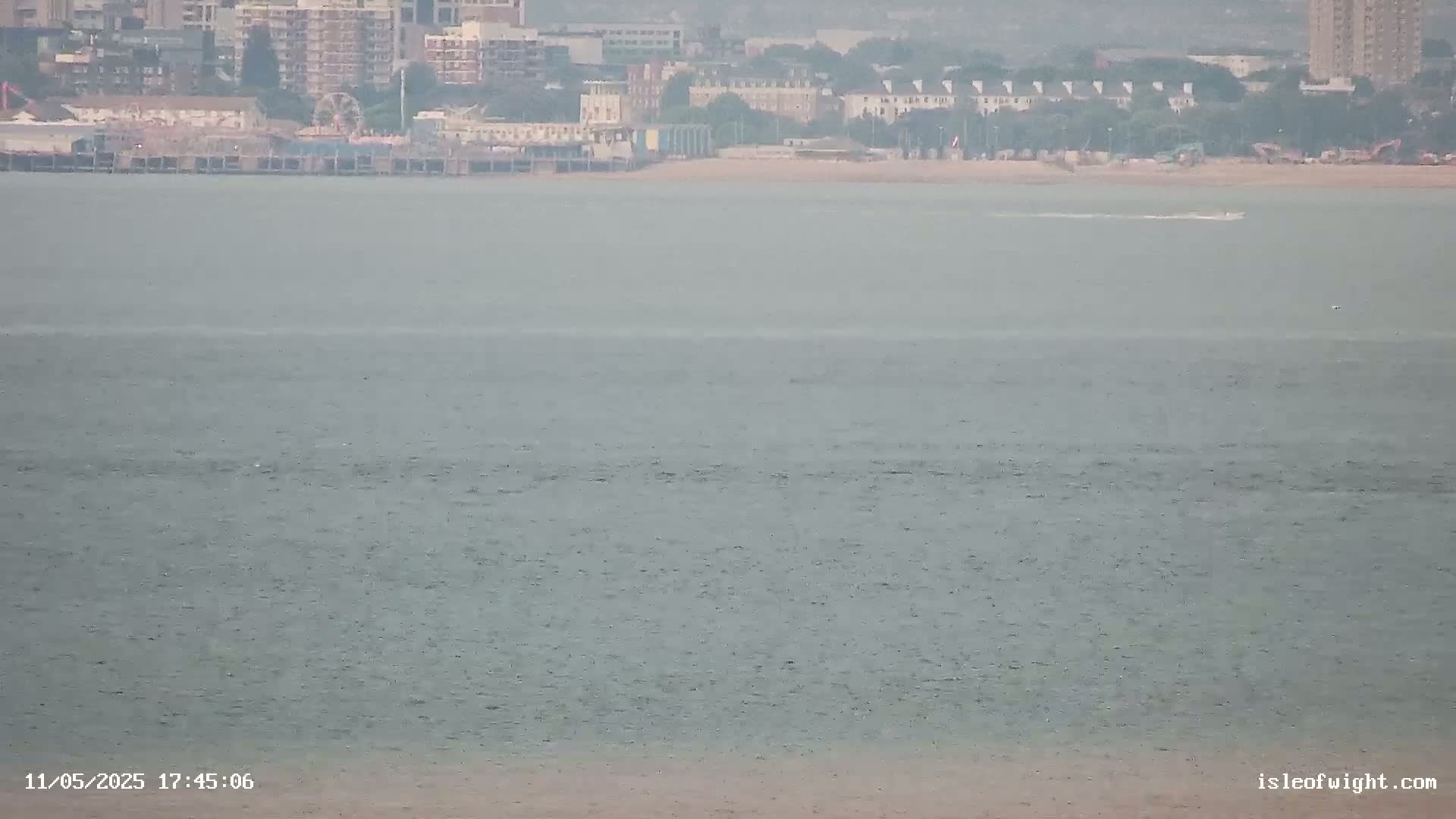 A hazy view shows a calm body of water with a distant shoreline featuring buildings, a pier, and a beach.