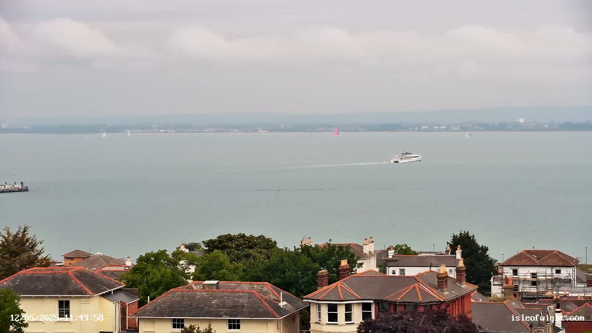 A ferry travels across a calm, gray body of water under an overcast sky, with a collection of houses and trees visible in the foreground.