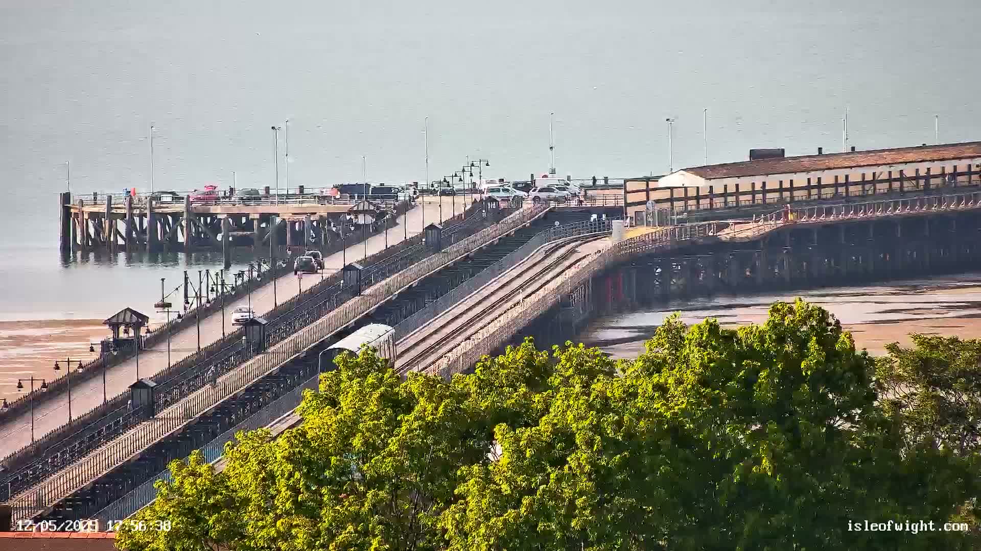 A long pier extends over calm, shallow water, with a train traveling along its tracks and cars parked on the pier's roadway.