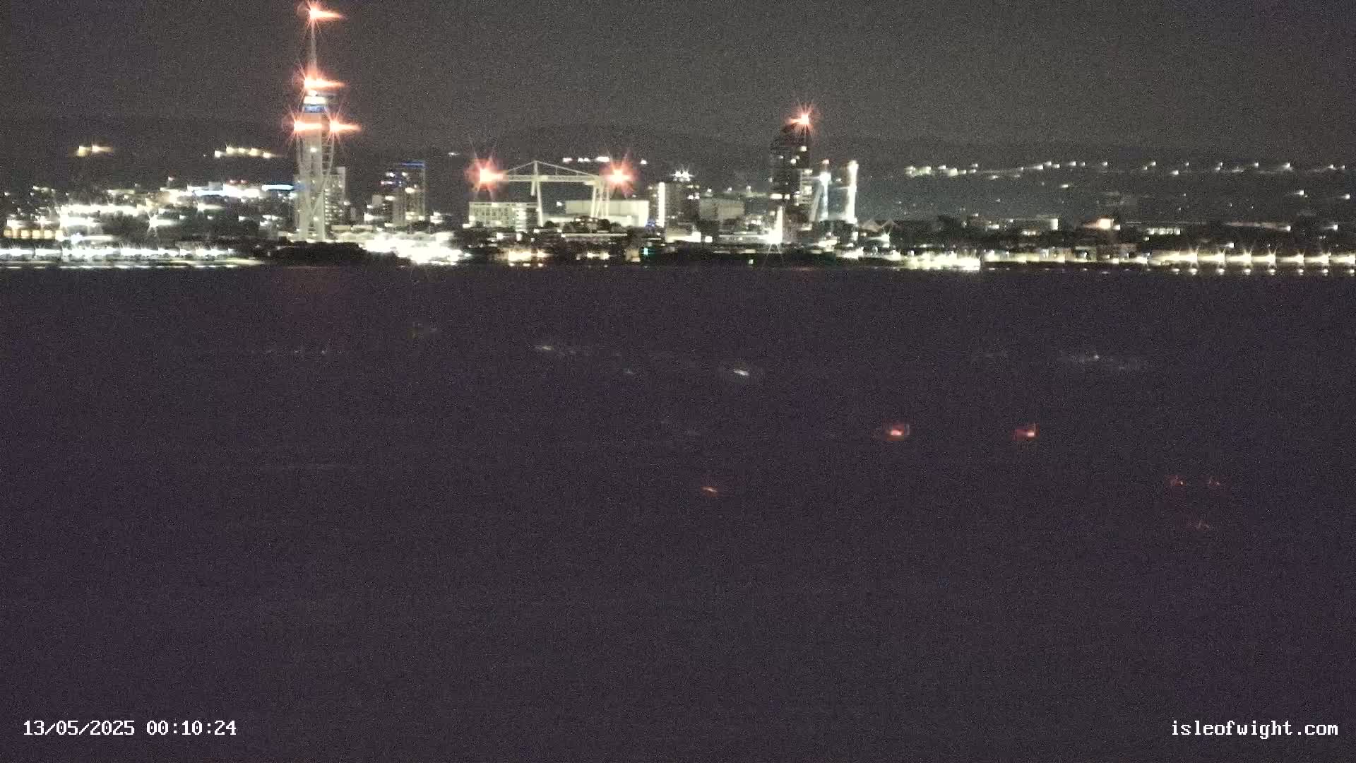 A nighttime view of a city skyline across a dark body of water, illuminated by various lights.