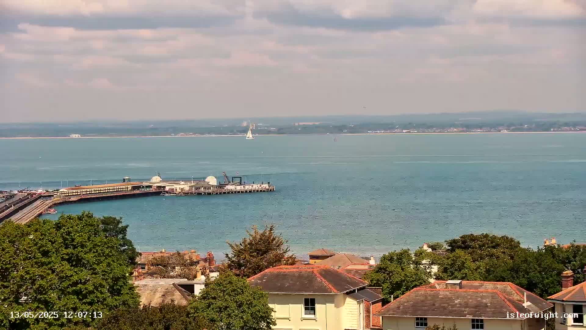 A seaside town with houses and green trees overlooks a calm, blue body of water featuring a pier and a sailboat in the distance under a partly cloudy sky.