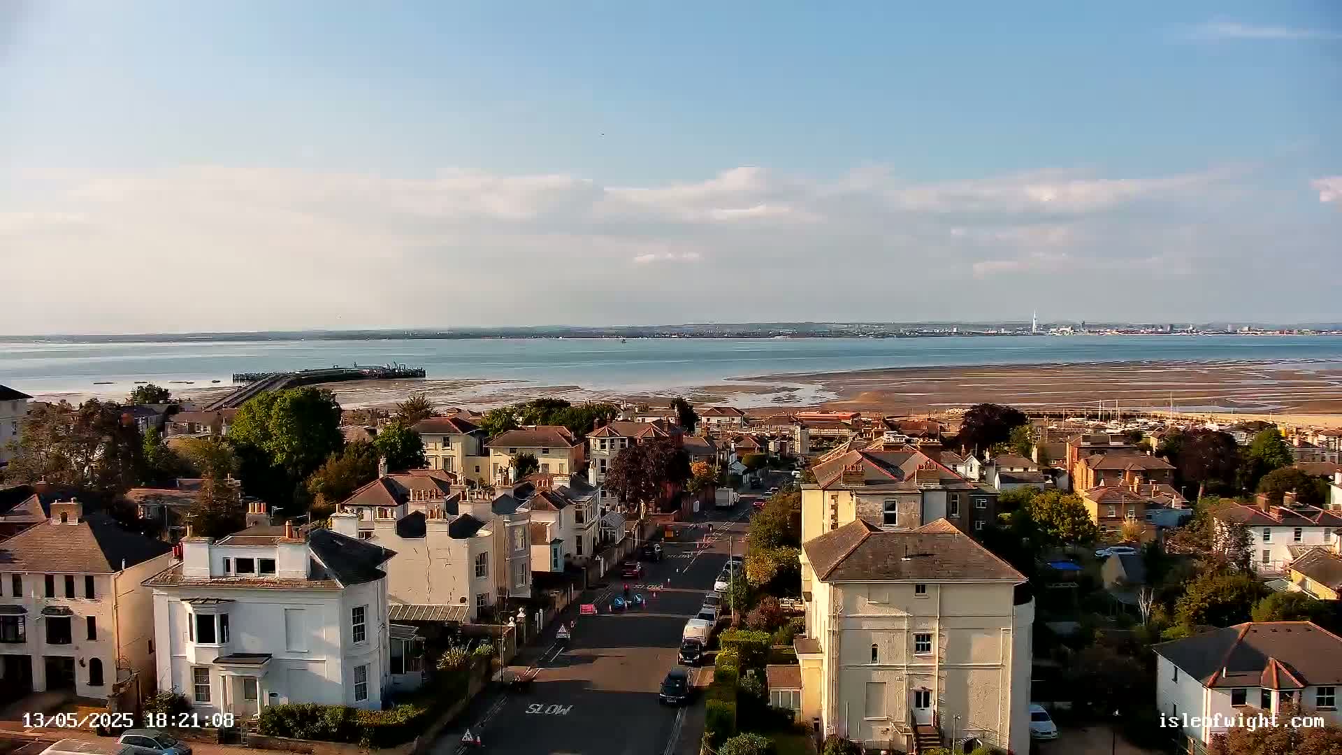 A coastal town with two-story houses, a road, and a body of water under a partly cloudy sky.