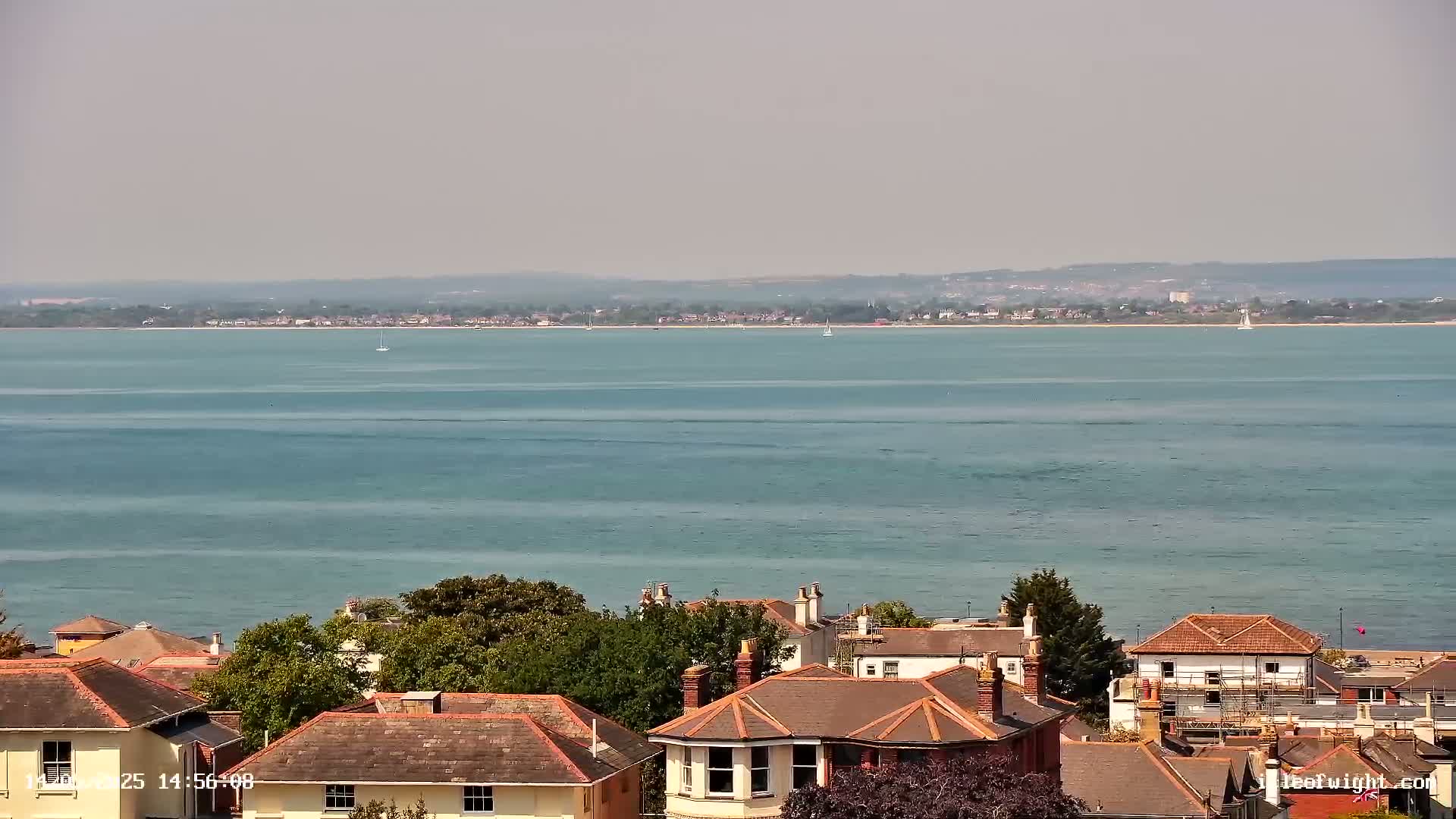 A calm, light-blue body of water stretches to a hazy, distant shoreline, beyond which sits a collection of houses with red-tiled roofs nestled amongst green trees.