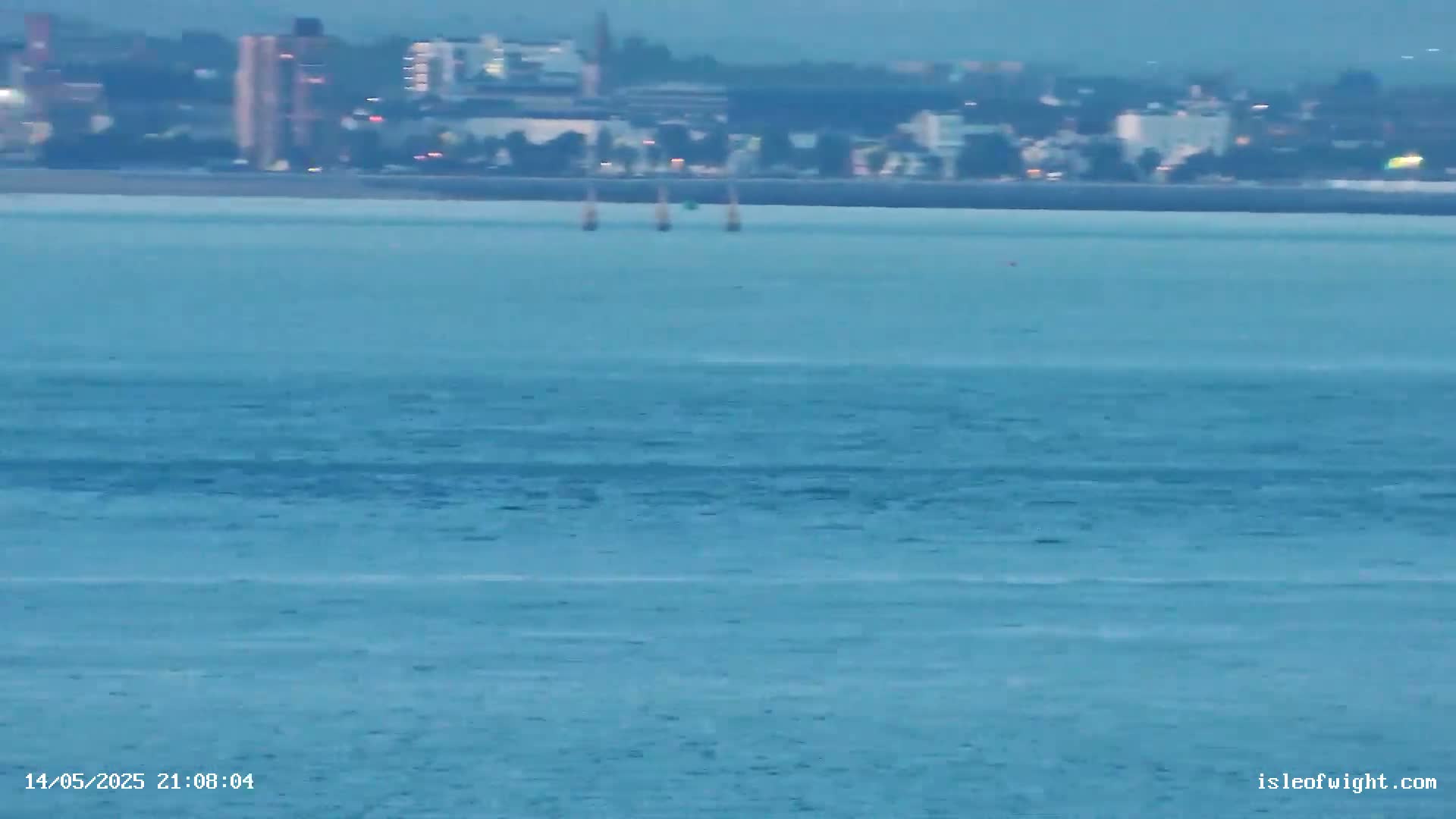 Three sailboats are visible on a calm, blue body of water in front of a distant cityscape at twilight.
