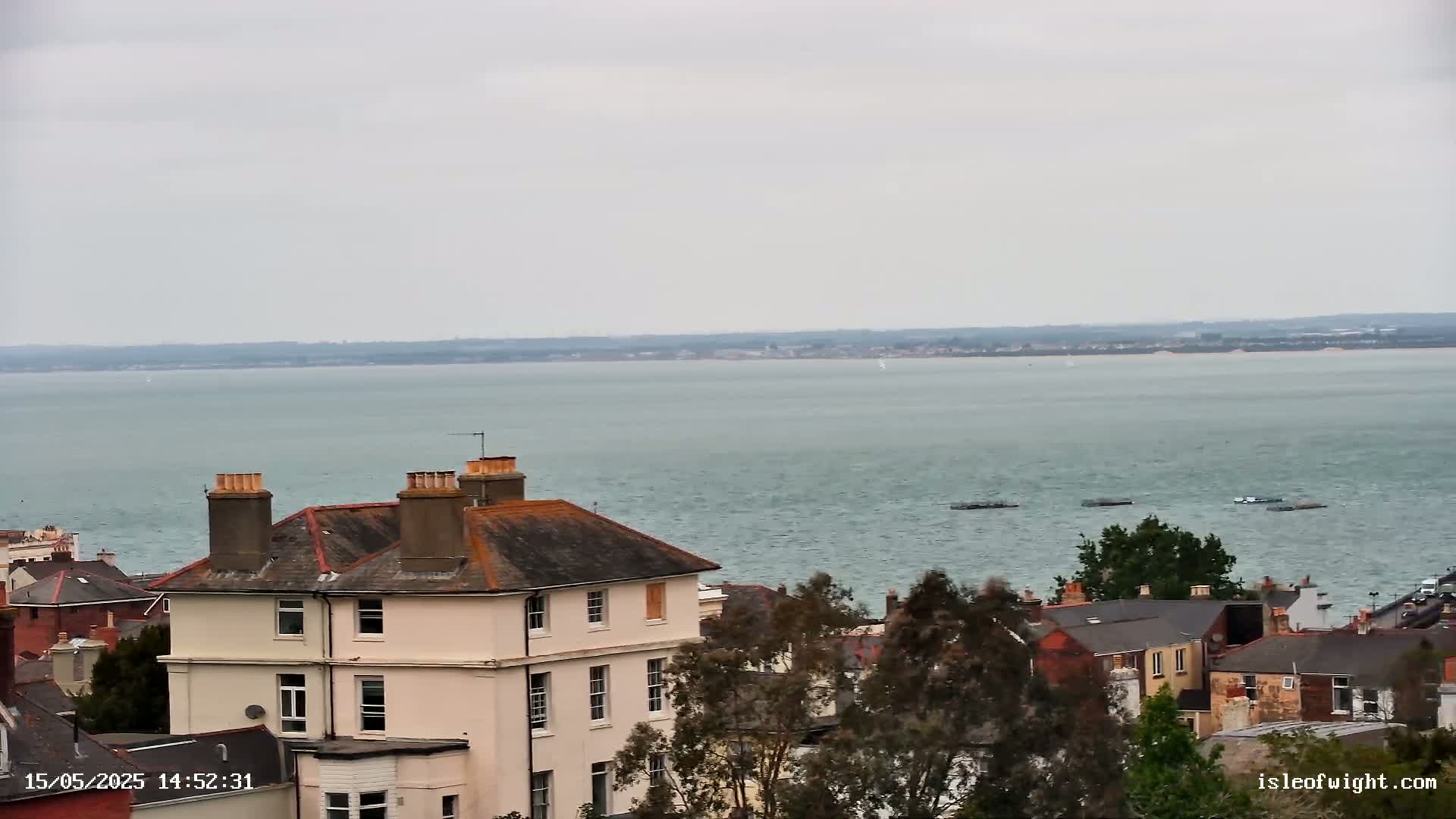 A three-story light beige building with a brown roof is situated among other buildings and green trees overlooking a calm, grey-green body of water under an overcast sky.