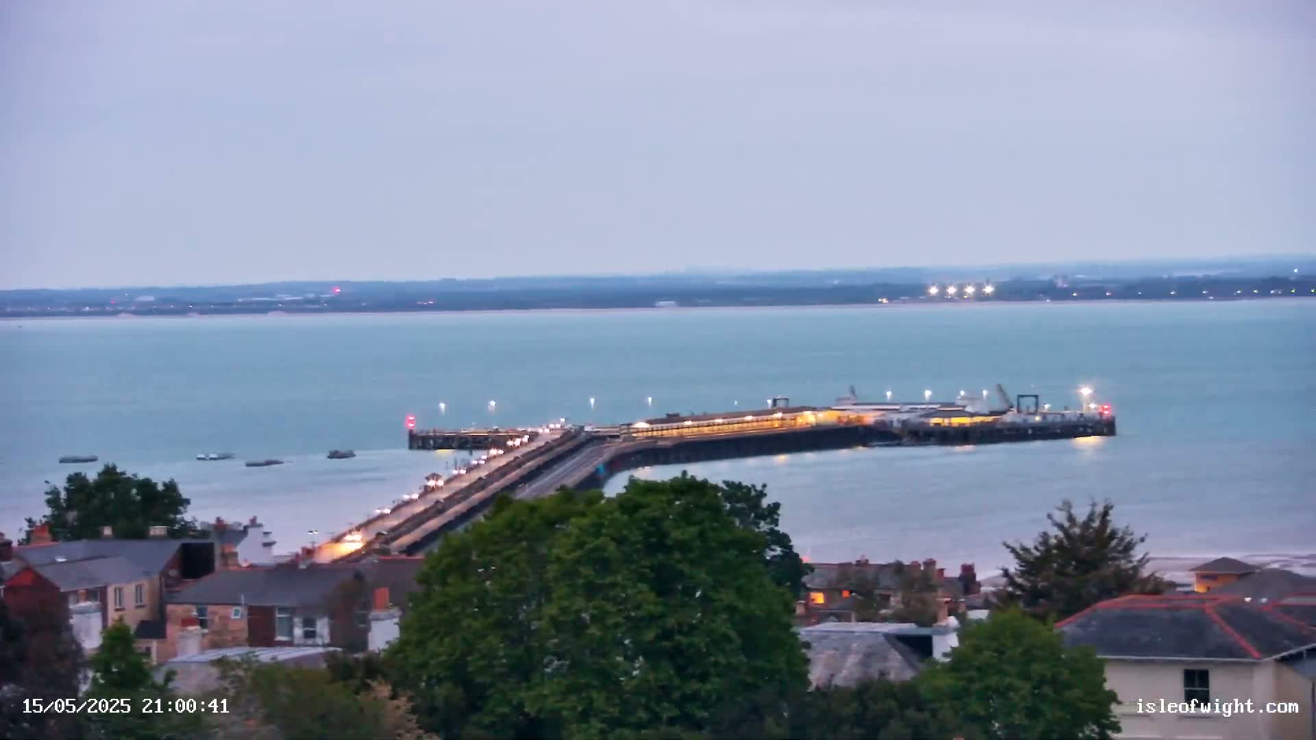 A brightly lit ferry terminal extends into a calm, twilight-colored sea, with a town visible in the foreground.