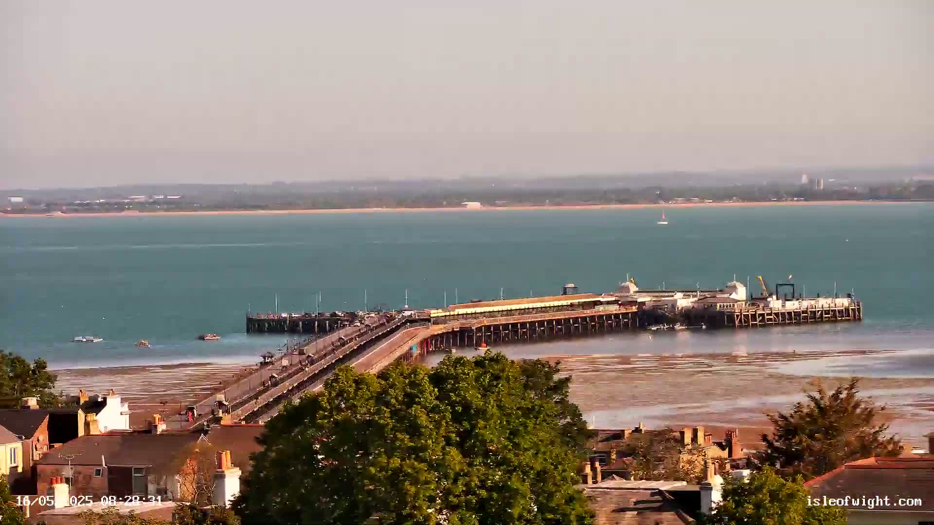 A long pier extends from a shoreline into a calm, teal-colored body of water, with a low-tide exposing a wide expanse of sand, and small boats visible in the distance.
