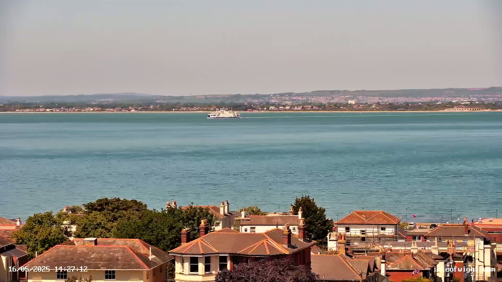 A ferry crosses a calm, blue body of water in the distance, while in the foreground, red-tiled rooftops of houses are nestled amongst green trees.