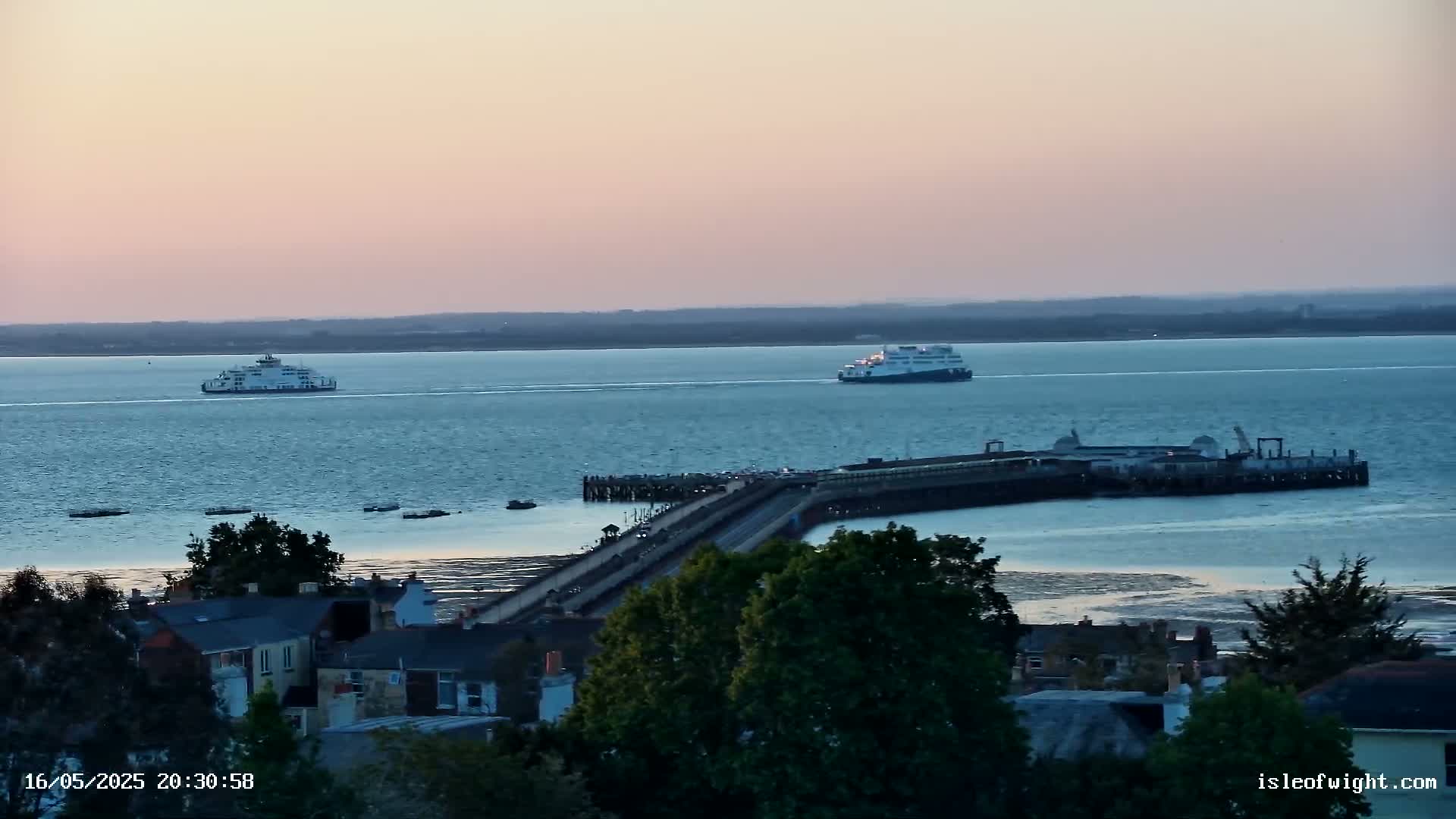 Two ferries are on a calm body of water at dusk, near a long pier that extends from a town's edge.