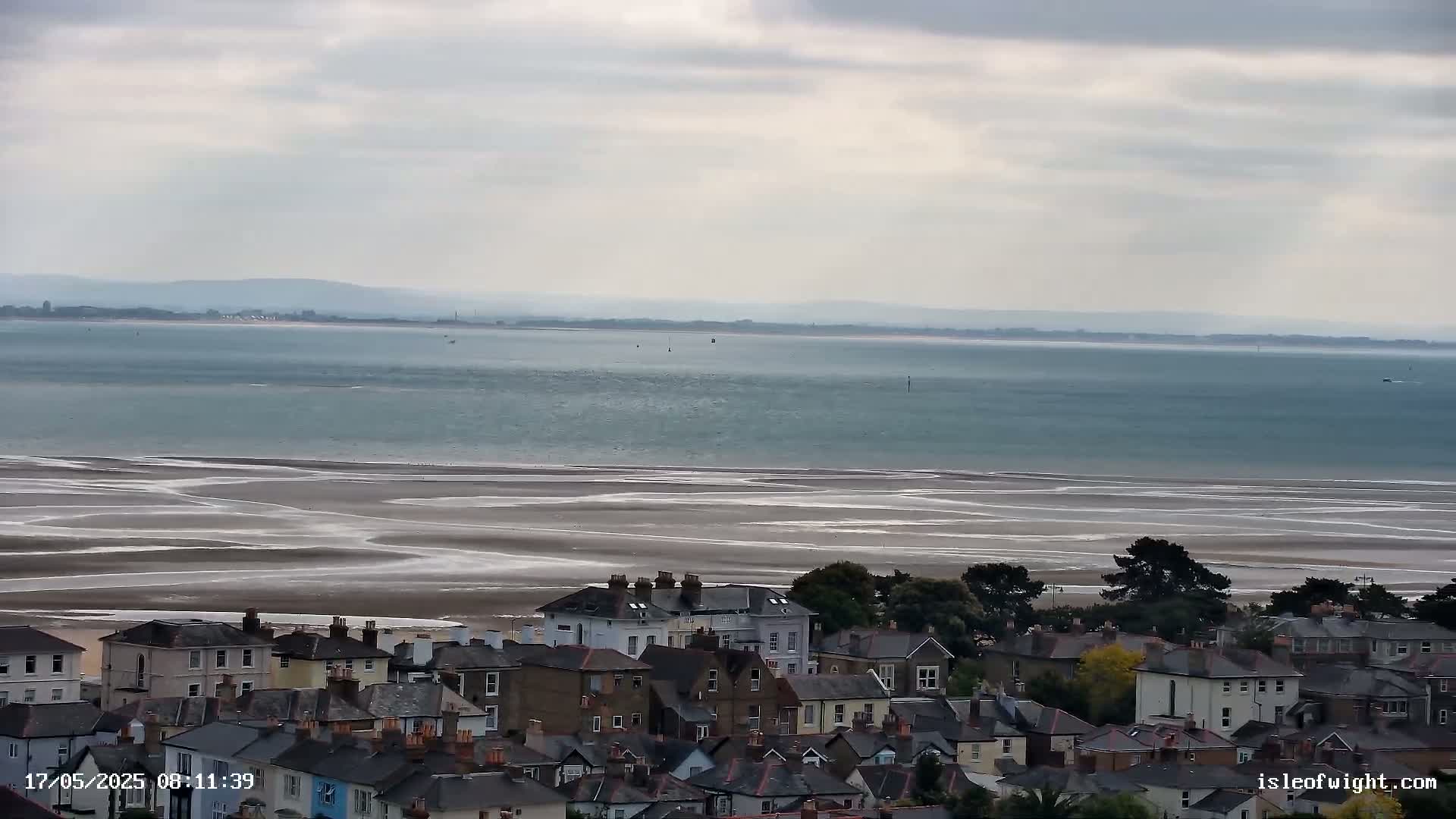 A coastal town overlooks a wide expanse of calm, light-blue water and a broad, sandy beach patterned with receding tide lines under an overcast sky.