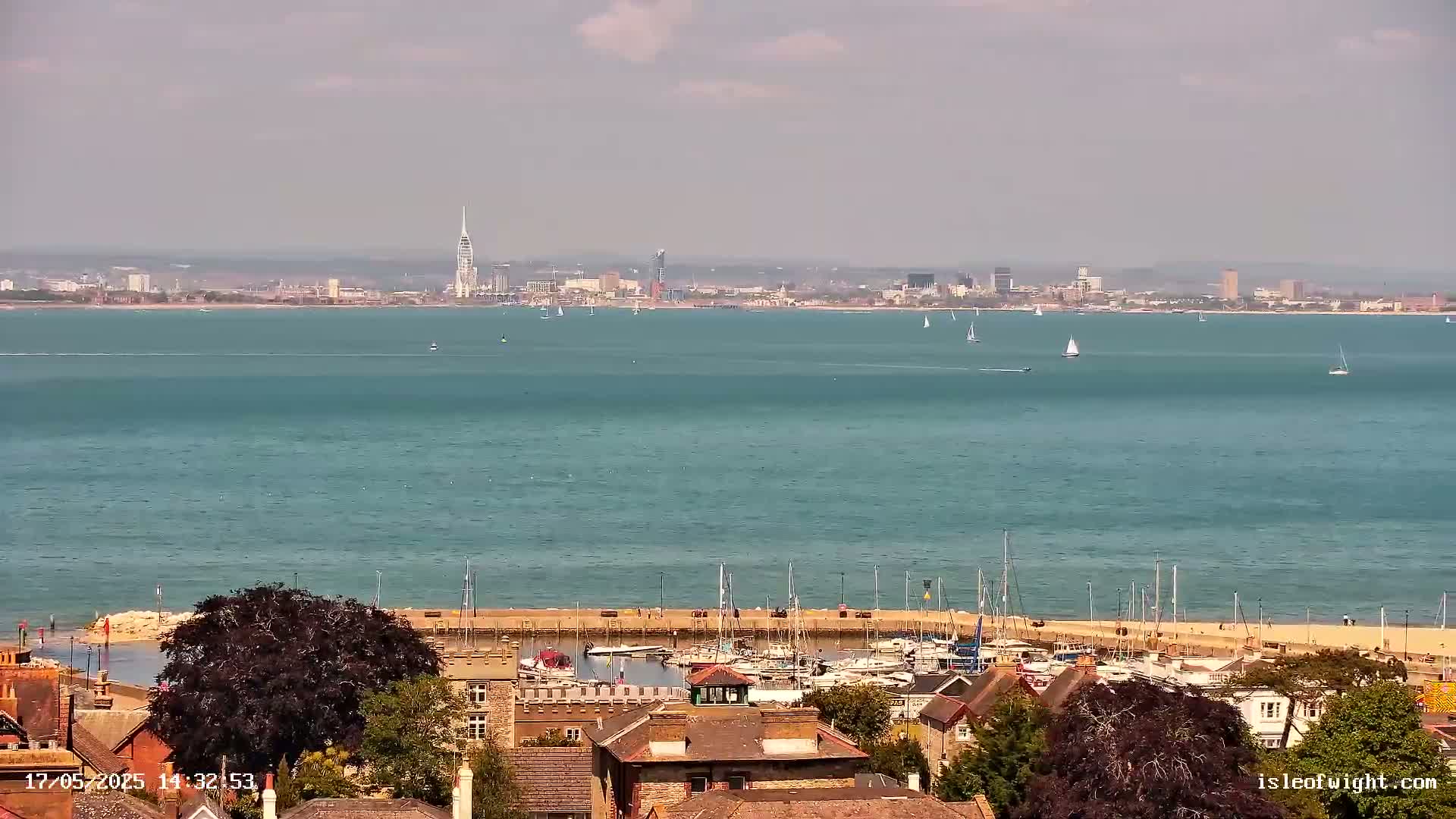 A hazy, sunny day shows a view of a harbor filled with boats, a shoreline with buildings, and a city skyline across a body of water.