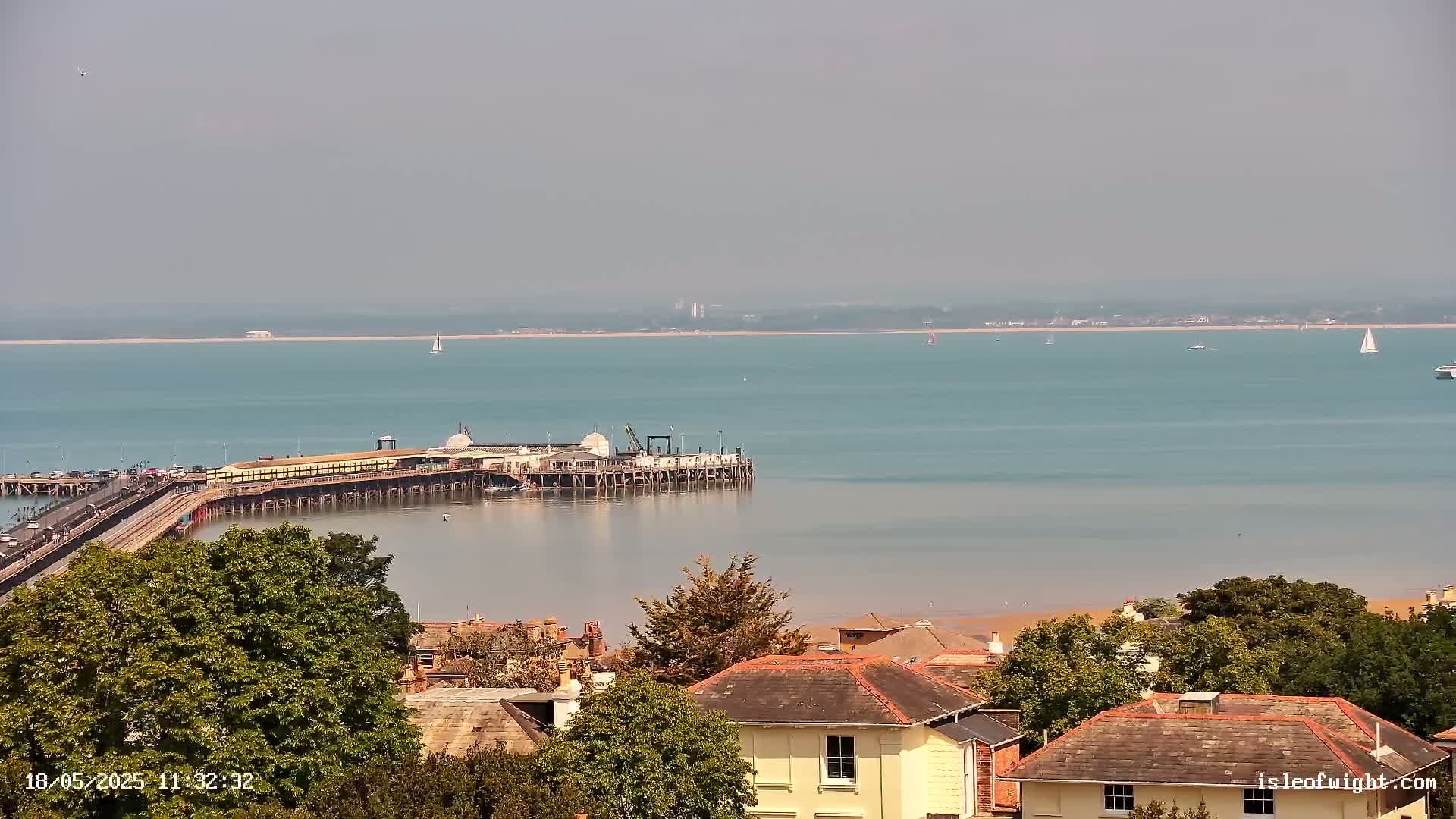 A long pier extends into a calm, light-blue sea under a hazy sky, with a town and green trees in the foreground.
